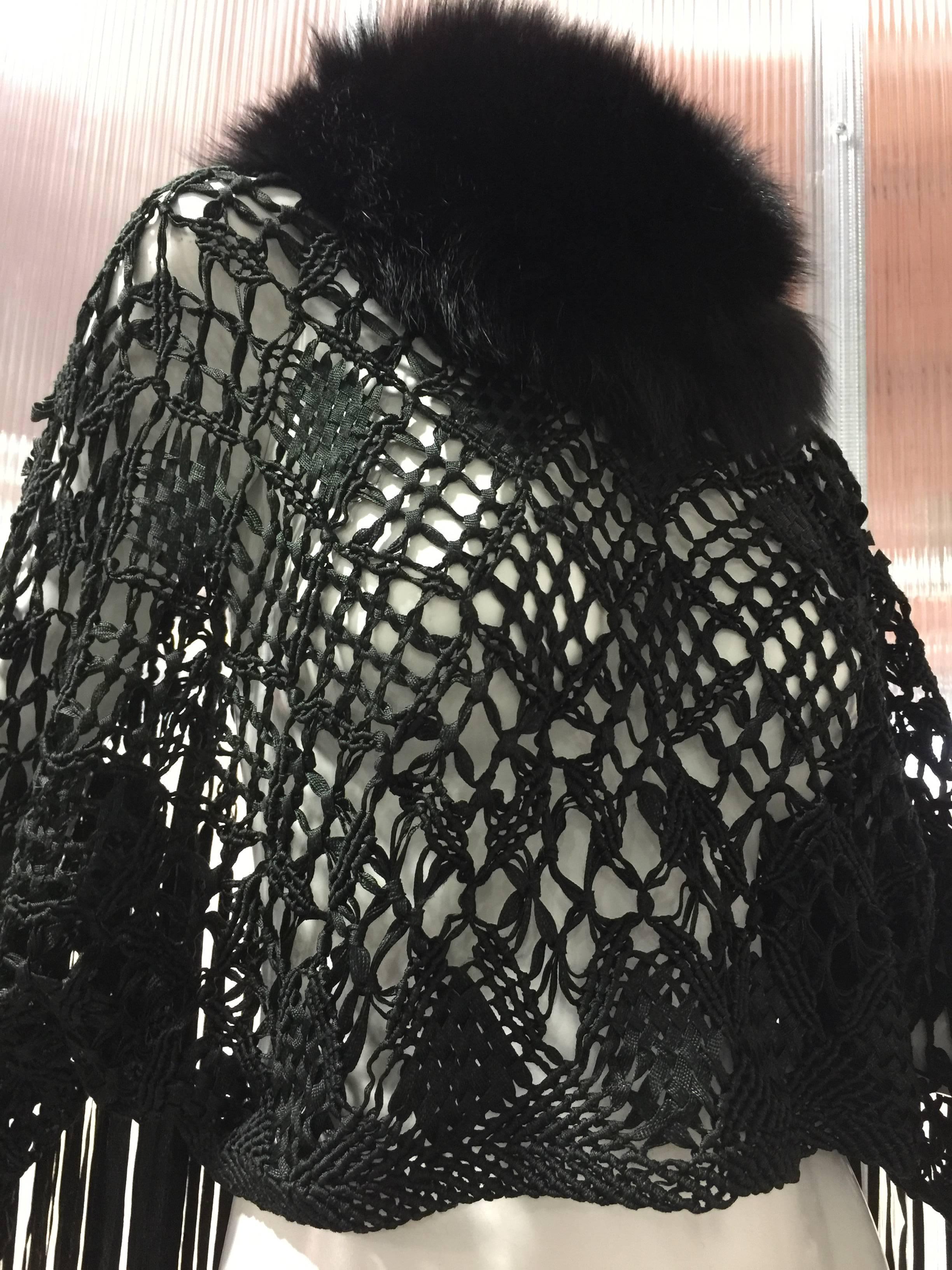 1970s black silk ribbon macrame and fringed cropped poncho is made to slip over the head and arms. Fluffy black fox fur tightly surrounds the neck for warmth and arms slide thru the poncho for movement allowing the fringe to sway about freely. A