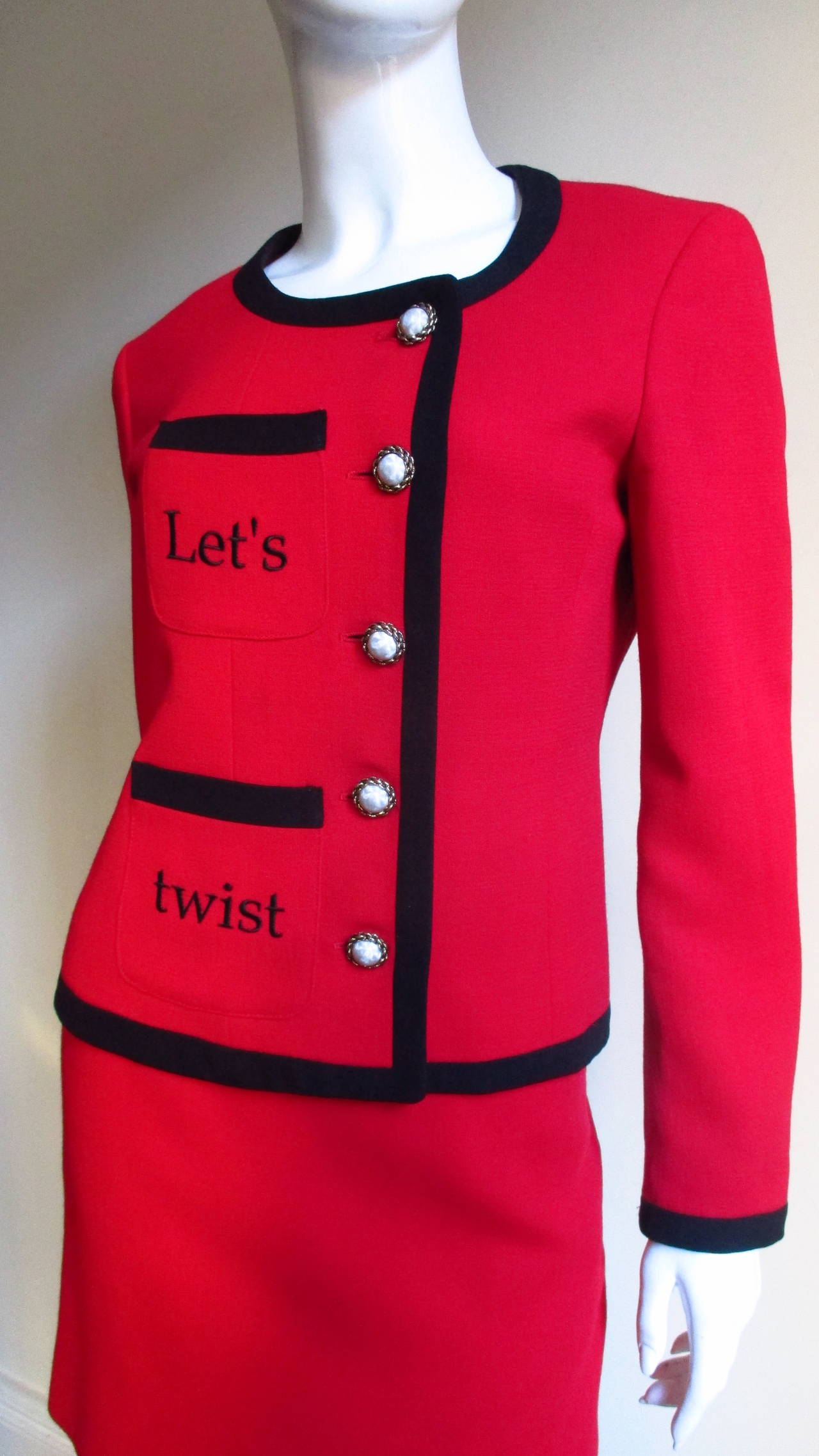 A fabulous red light weight wool suit from Moschino. The simple jacket closes off center with 5 faux pearl buttons.  The other side has 2 pockets one above the other with the words 'Let's' and 'twist' embroidered in black on each.  The back has the