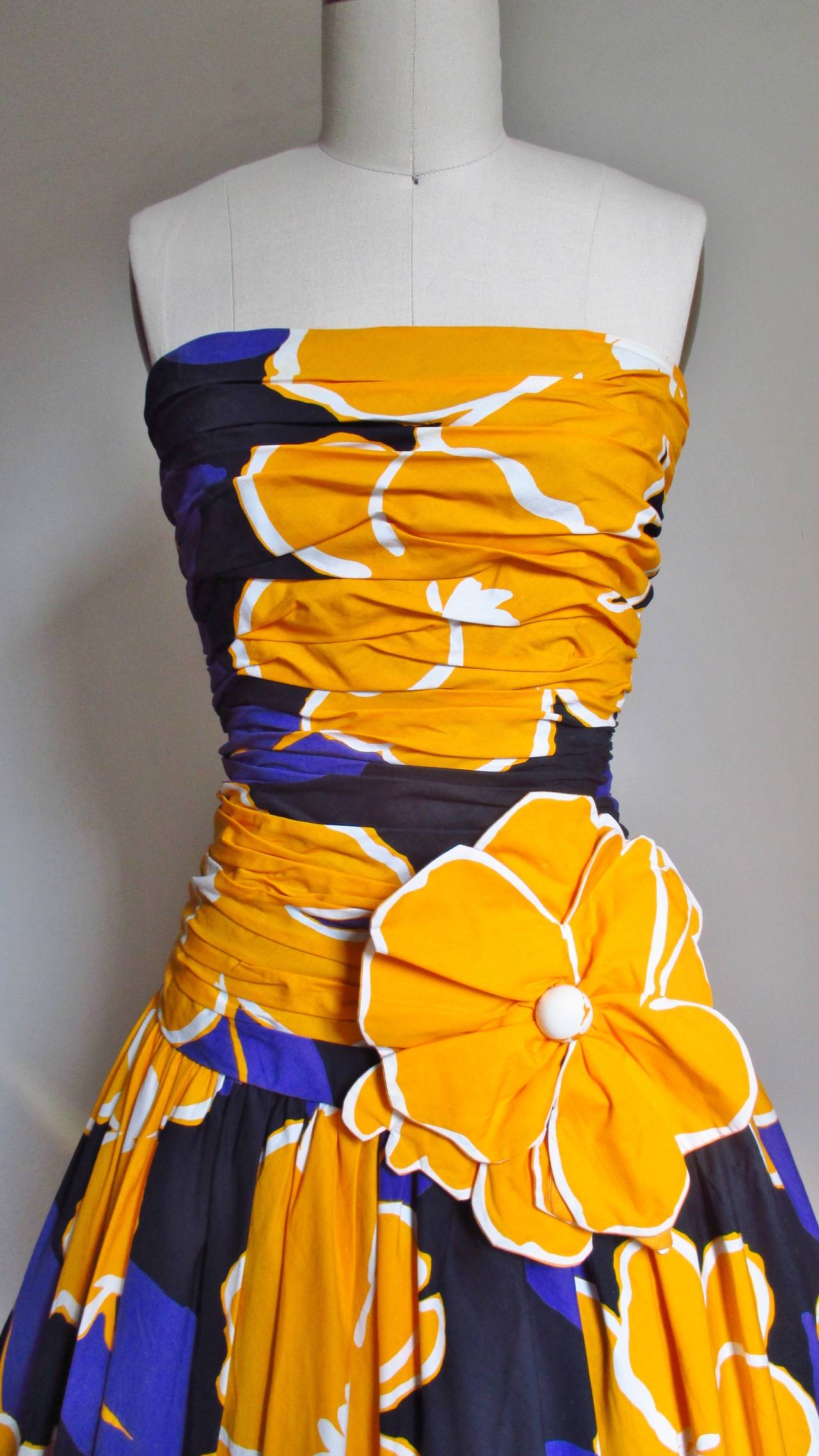 A very flattering bustier style cotton midi length dress dress from Mignon in purple and gold bold flowers on a black background.  It has a dropped waist ruched boned bodice with a full gathered skirt and a large applique flower off center front