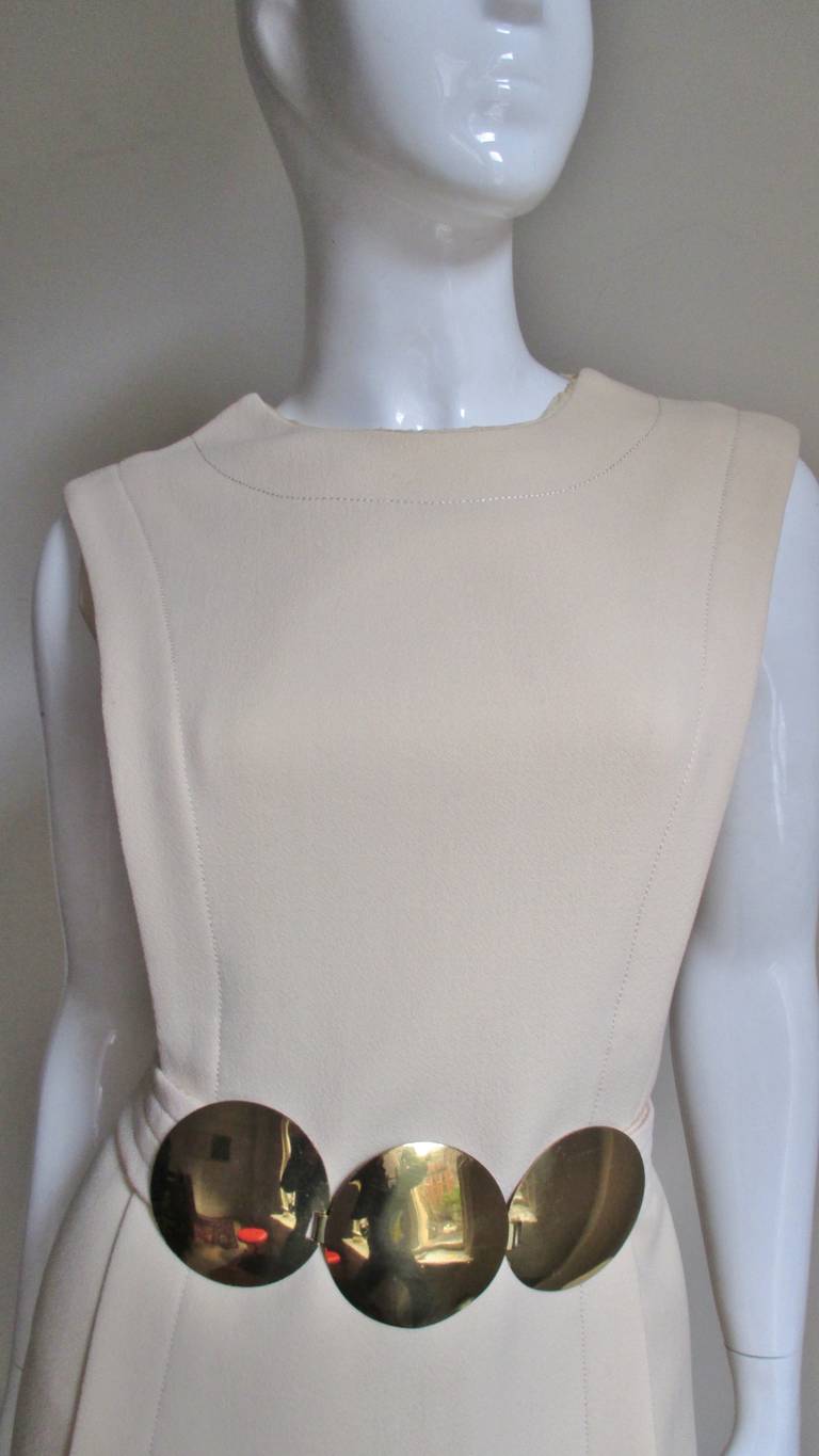 A fabulous off white wool crepe dress from Pierre Cardin.  A simple A line shift with an attached tabard which separates from the waist down front and back and is several inches shorter than the dress. The belt is incredible- 3 3" gold discs