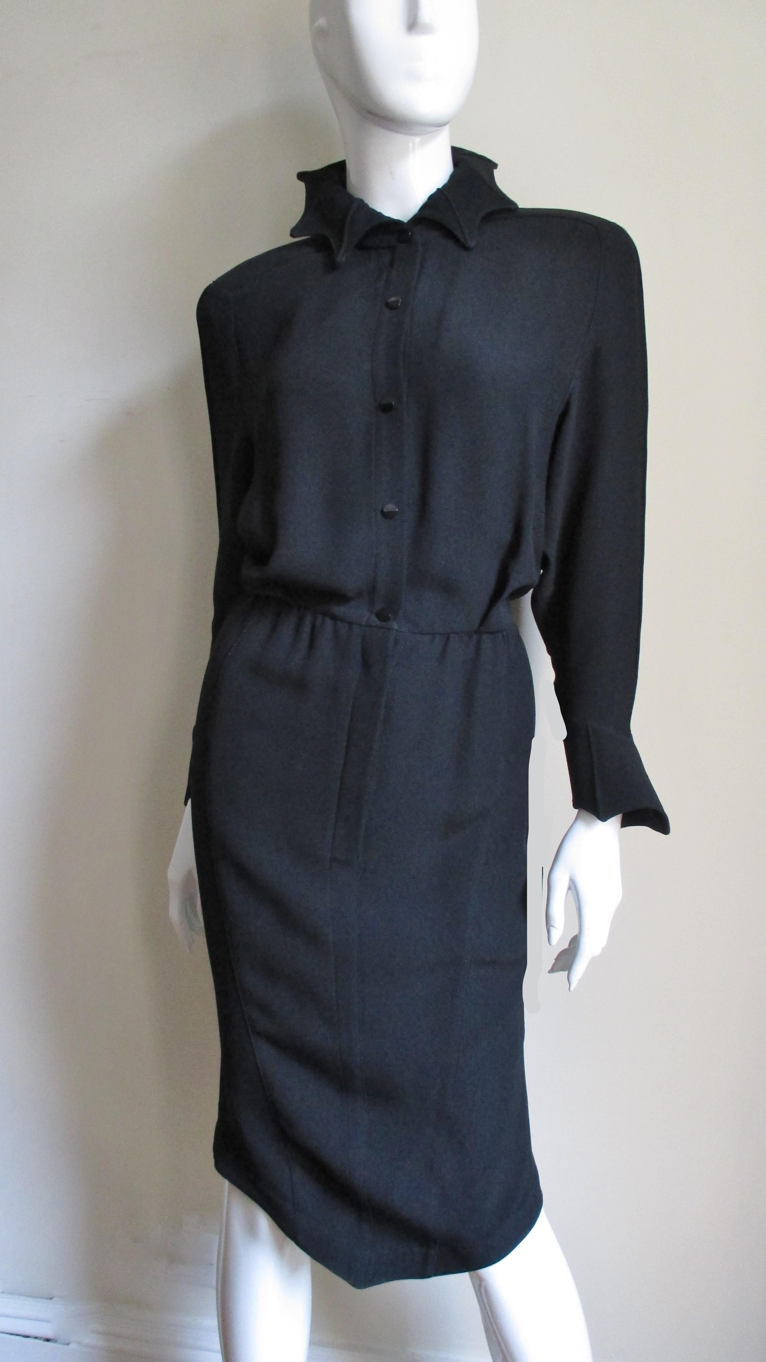 A sort of modern Gothic twist on the wiggle dress from master designer Thierry Mugler in a rich black synthetic blend.  It has long cuffed sleeves, a stand up collar (can be worn up or down) and a blouson bodice with 3 tucks at the waist.  The cuffs