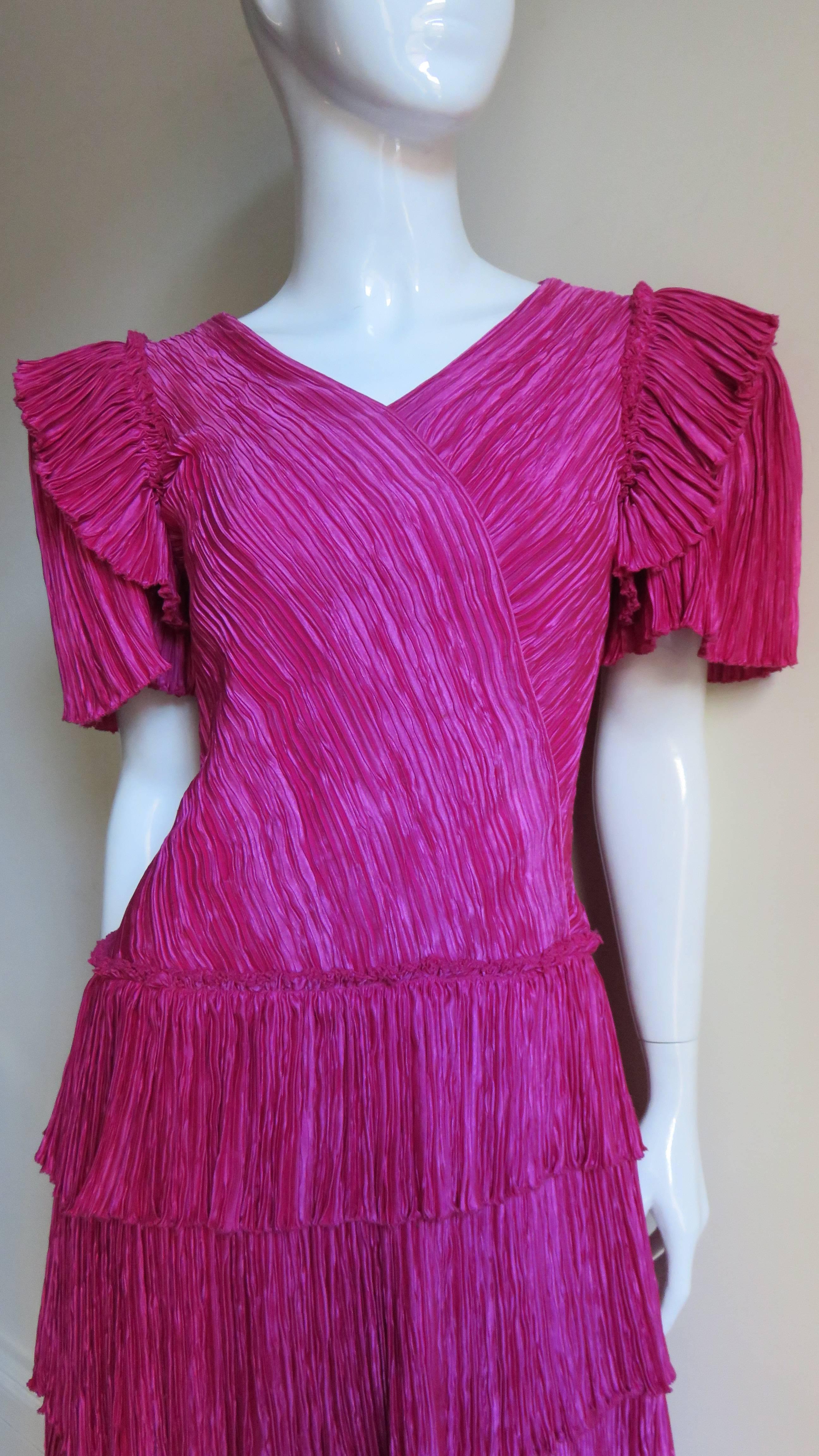 Une jolie robe micro plissée en soie rose de la ligne Couture de Mary McFadden.  Elle a une encolure en V, un effet enveloppant sur le corsage devant et derrière et les manches courtes ont un volant autour des épaules.  La jupe A line se compose de