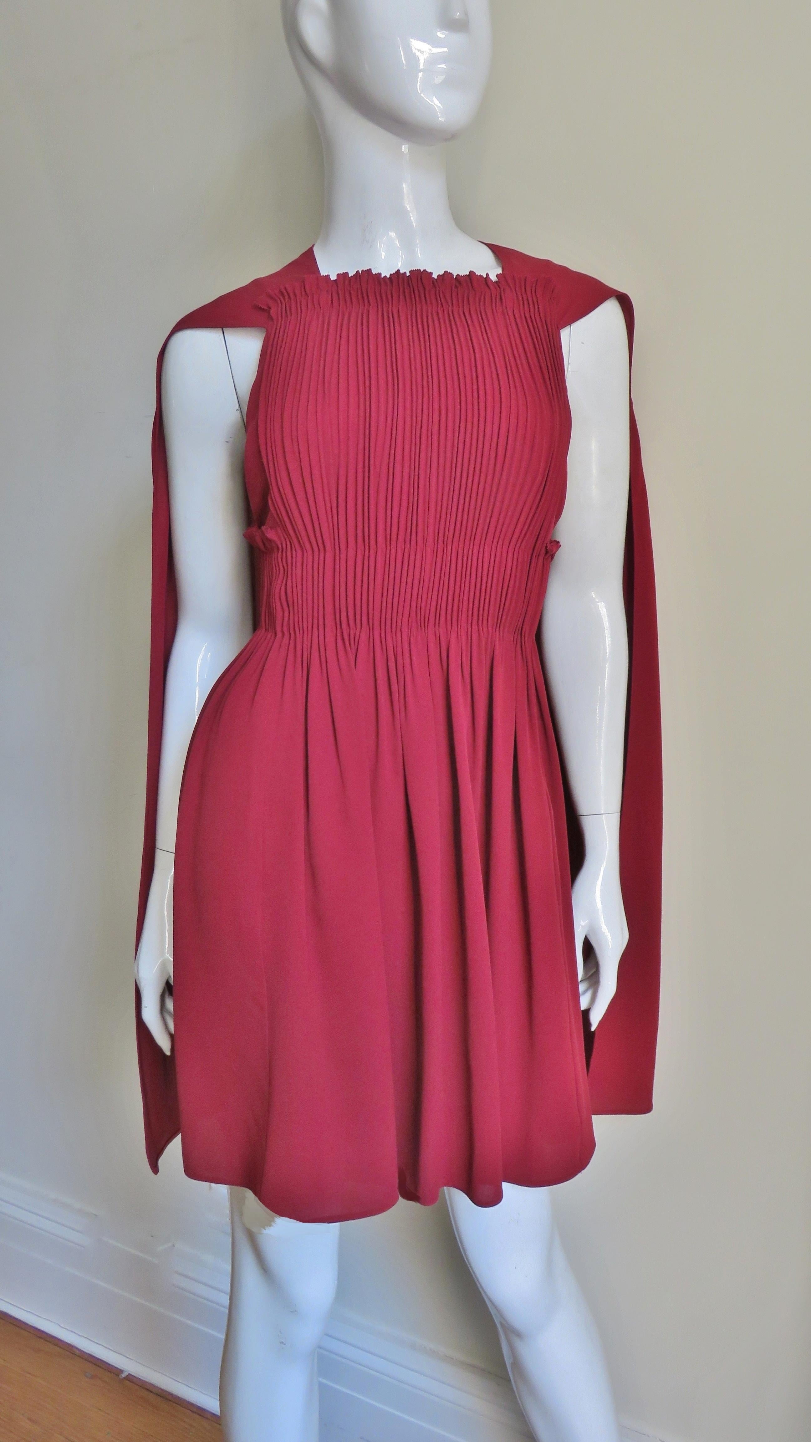 An incredible deep red dress with cape from Valentino. It has a vertically ruched front bodice (with straps in the back) that releases at the high waist into a full skirt.The underarms are deeply cut and a cape emanates from the shoulders. The