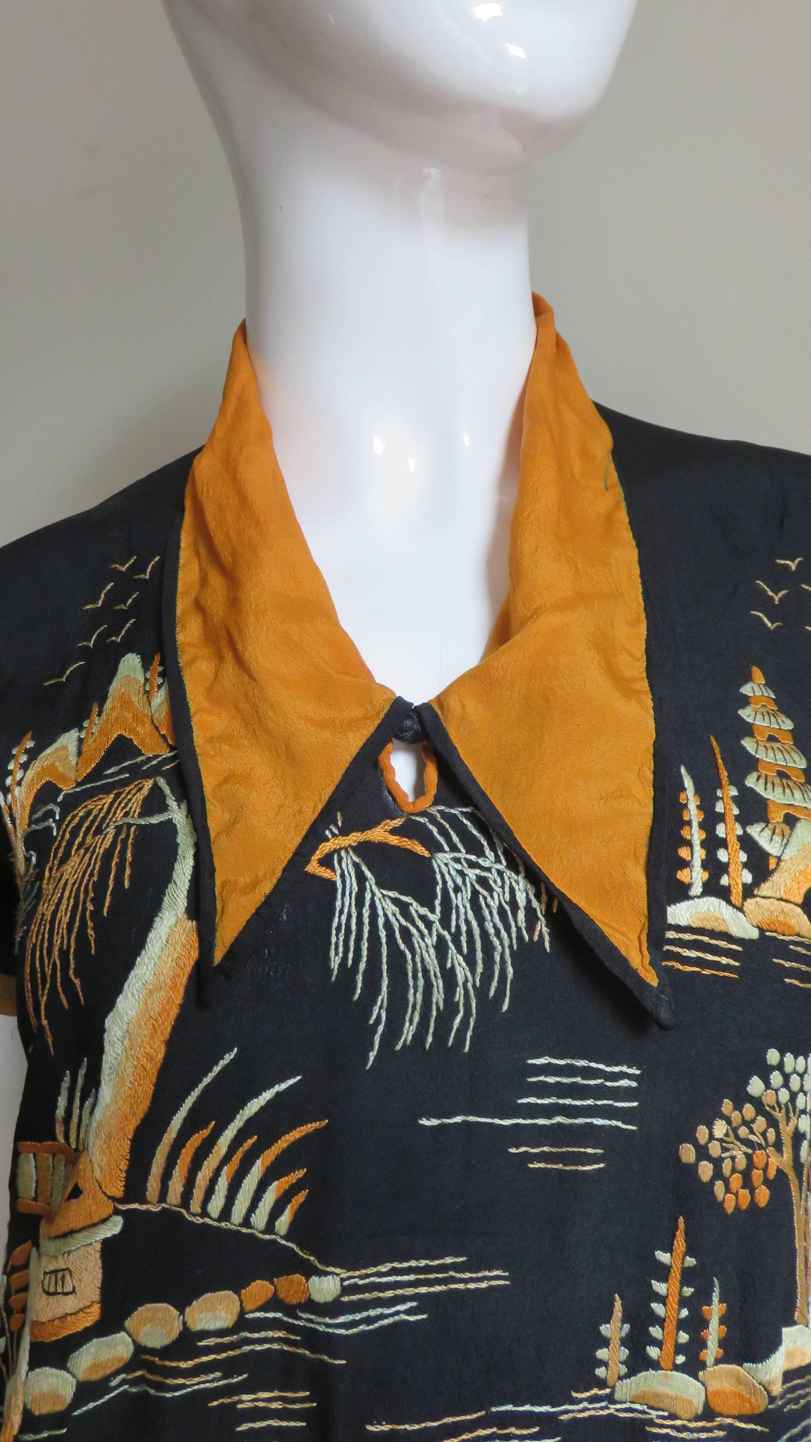 A pretty Asian embroidered black blouse with orange trim and collar.  It is elaborately embroider on the front with mountains, trees, pagodas.  It has a pointed shirt collar with a keyhole opening below it closing with a delicate woven frog.  The