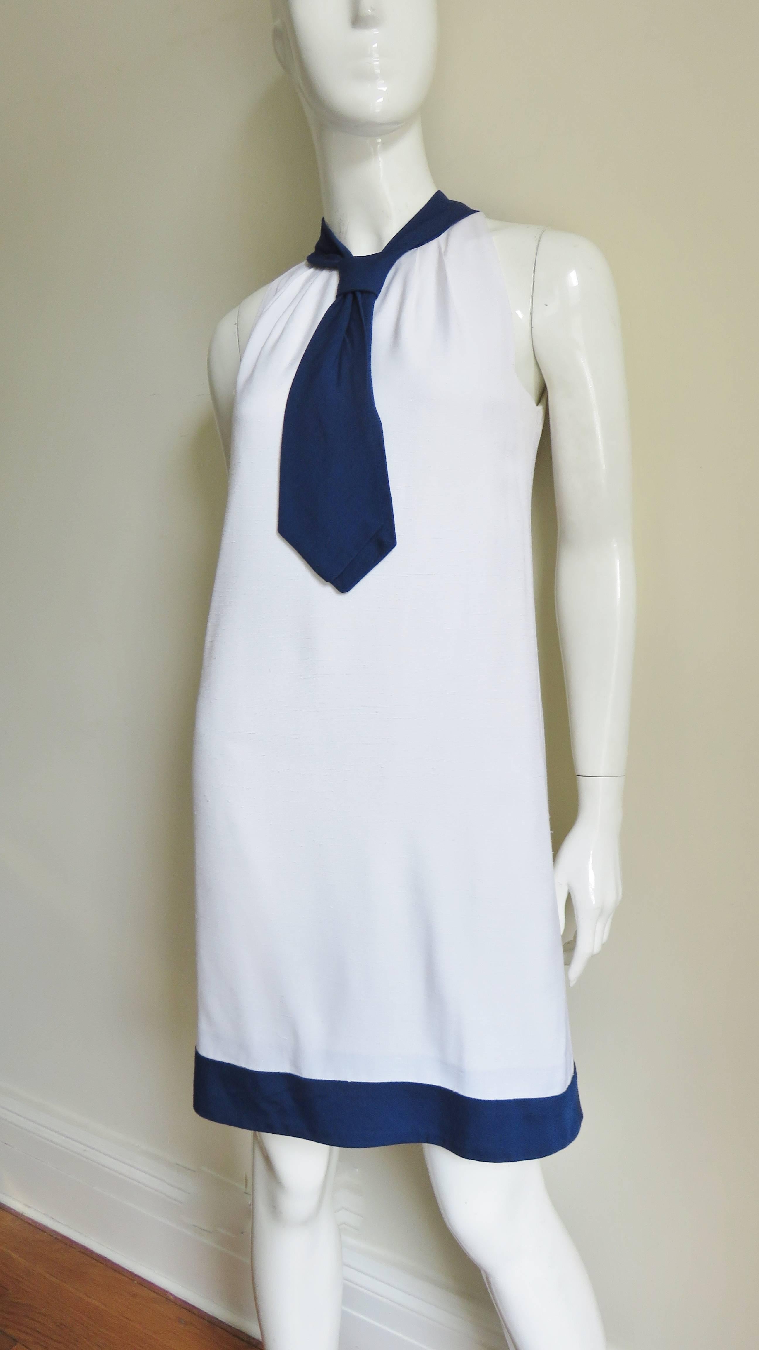 A great white and navy linen blend dress from Pierre Cardin. It is sleeveless with a band of navy around the hem and the neck forming a tie. The shoulders are cut in and the dress gathers slightly at the neck then flares subtly towards the hem.  It