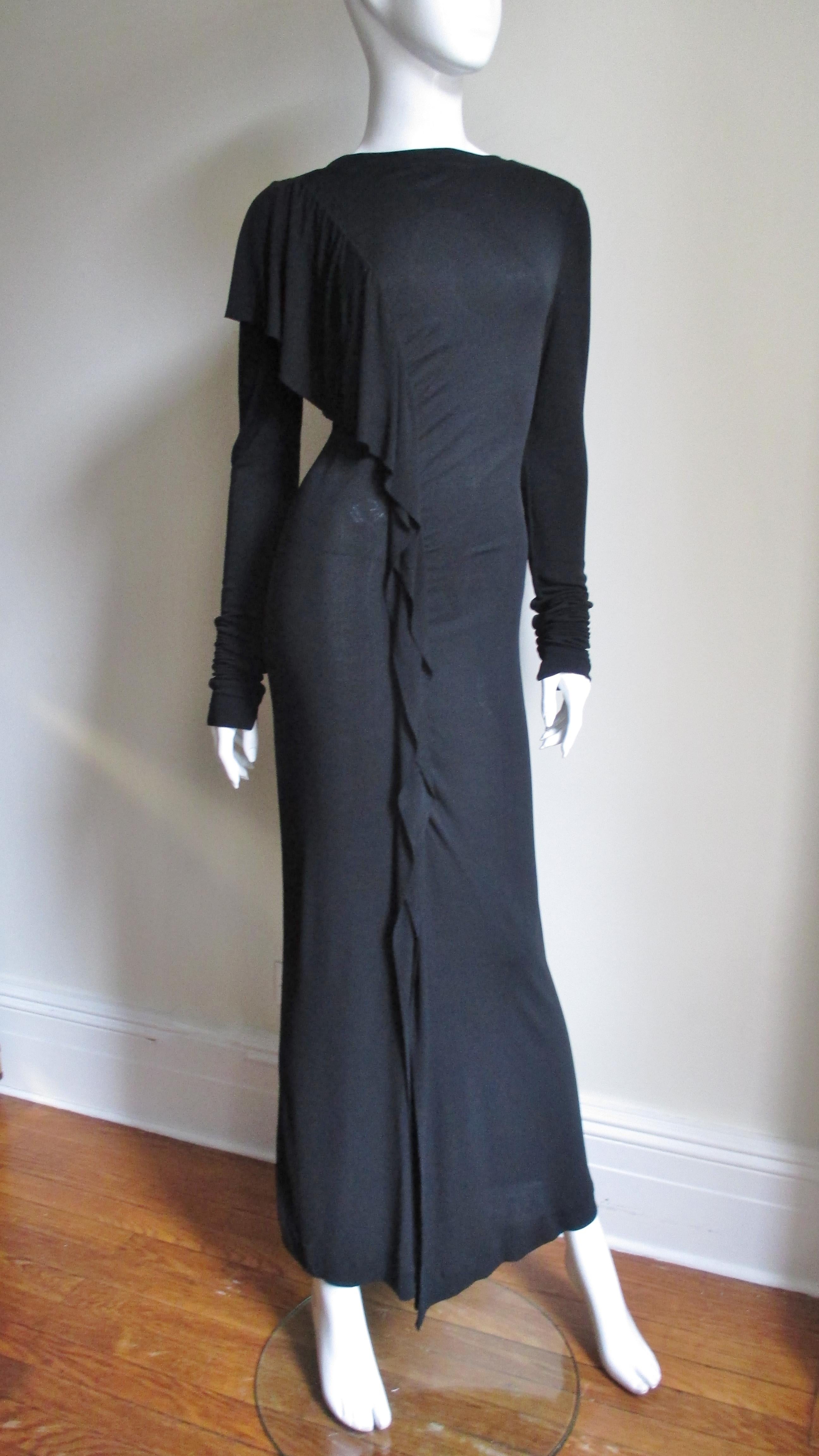 A fabulous black silk jersey dress from Jean Paul Gaultier.  It is semi fitted with a bateau neckline and a gradated draped ruffle wider at the front shoulder then narrowing to the hem which is open to the knee forming a front slit along that leg. 