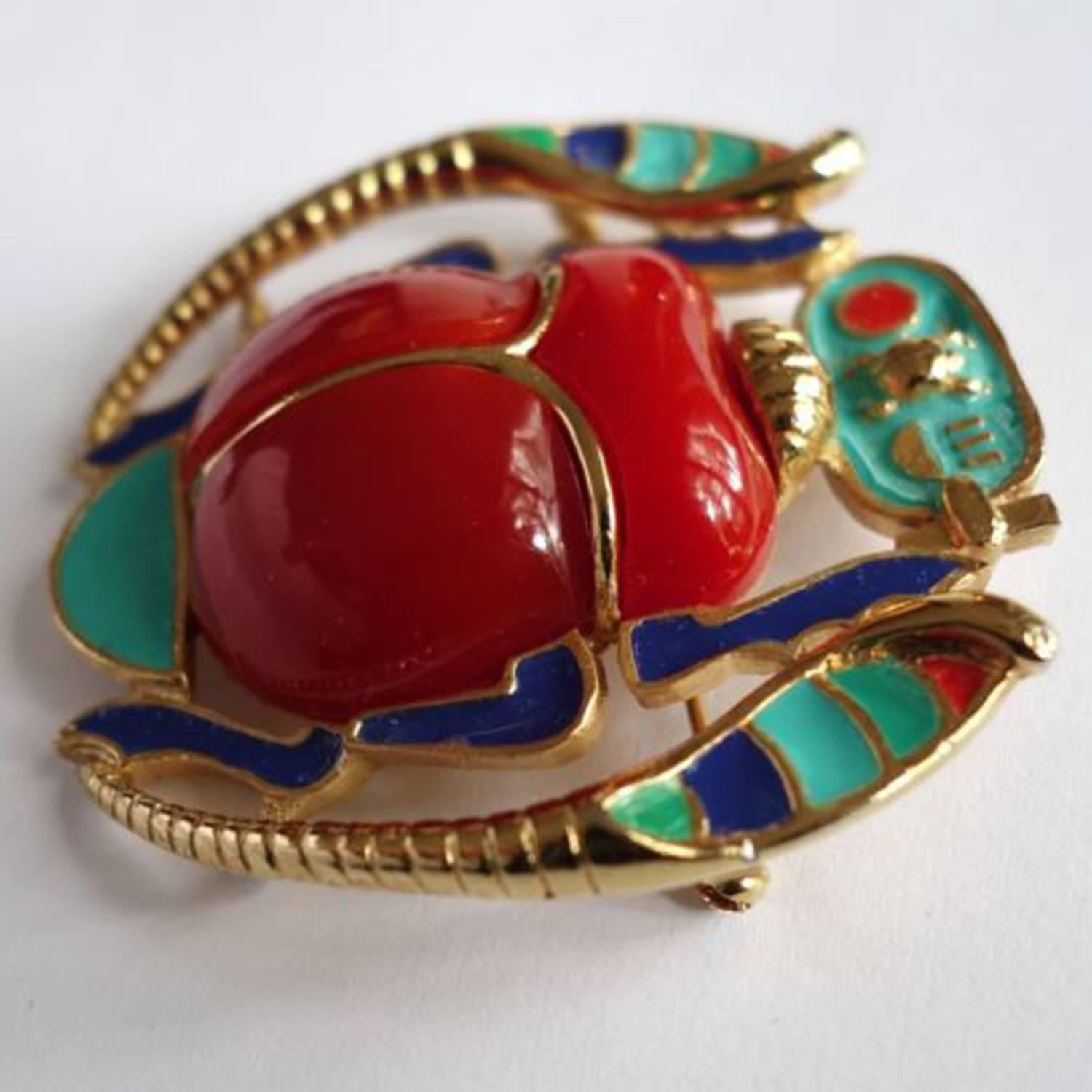 Featuring a vibrant red acrylic and enamel Scarab flanked by enamel accented stylized Serpents.  It measures approx 2.25" x 2.25"; signed: HATTIE CARNEGIE in oval cartouche on the underside. C1960s Chic and Timeless… Illuminating your Look