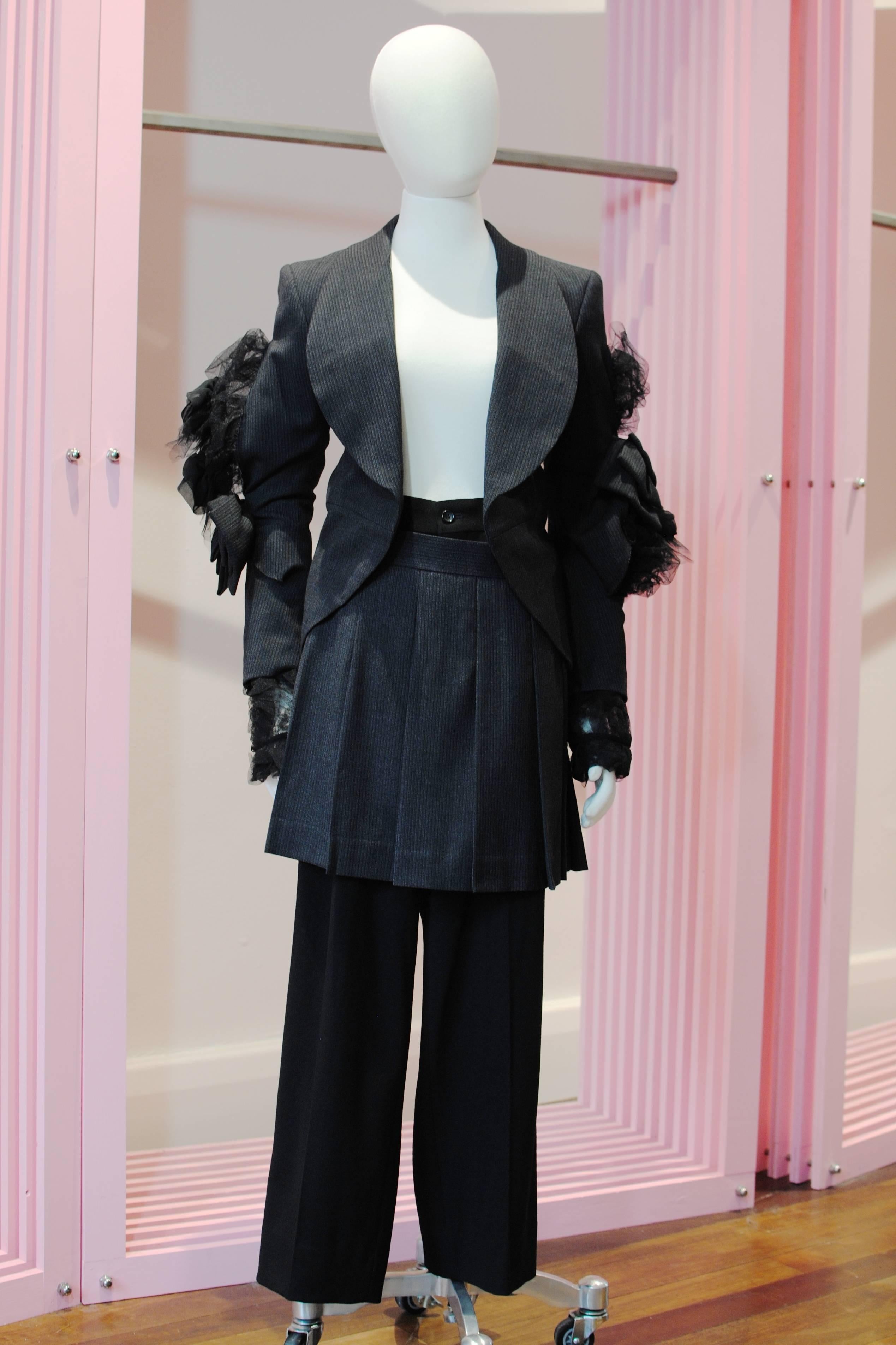 COMME des GARÇONS grey and black double layered suit from the 2004 Autumn/Winter collection. The jacket features a ruffled tulle under layer with attached rosettes which spills out through a dissection at the elbow that is patched up with a bow.