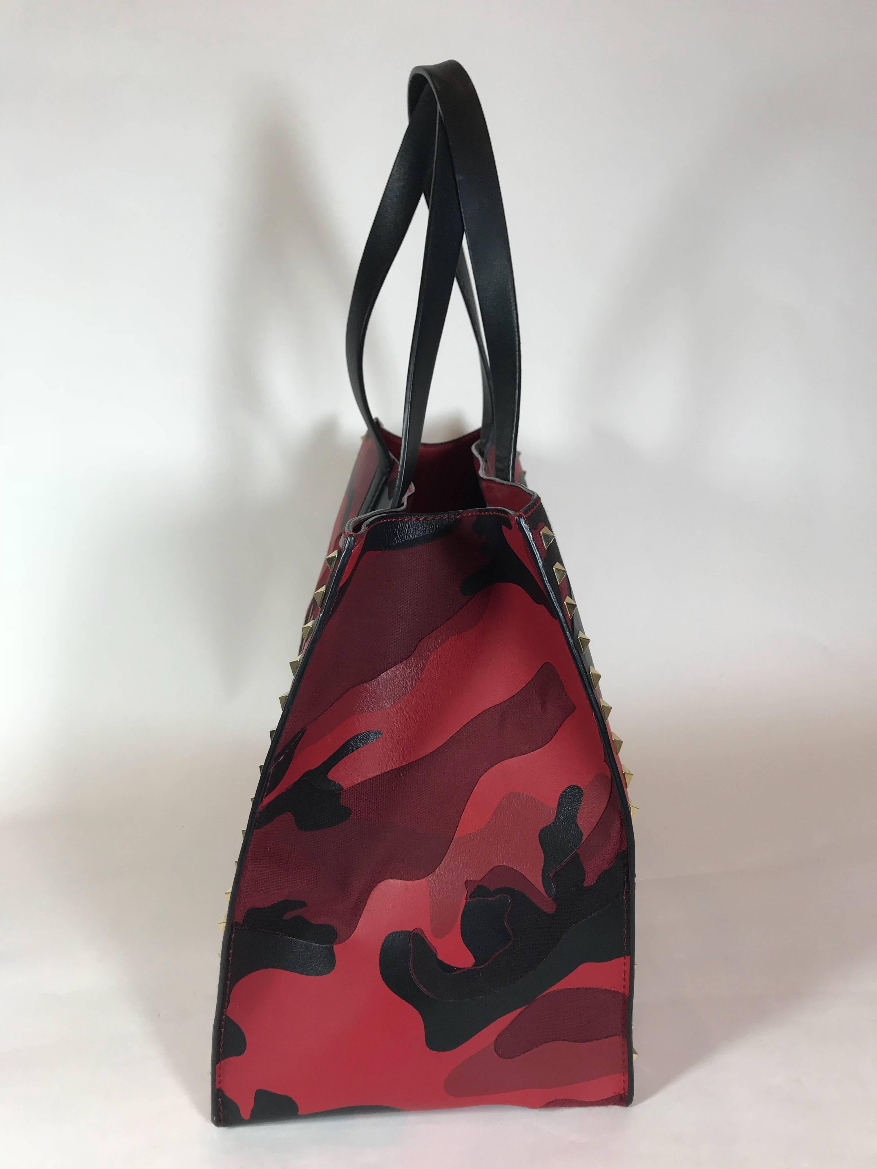 Red and black camouflage print canvas and leather. Gold-tone hardware. Magnetic snap closure at top. Dual flat shoulder straps. Black and tonal camouflage print leather trim. Rockstud stud embellishments throughout. Foil-stamped leather logo placard