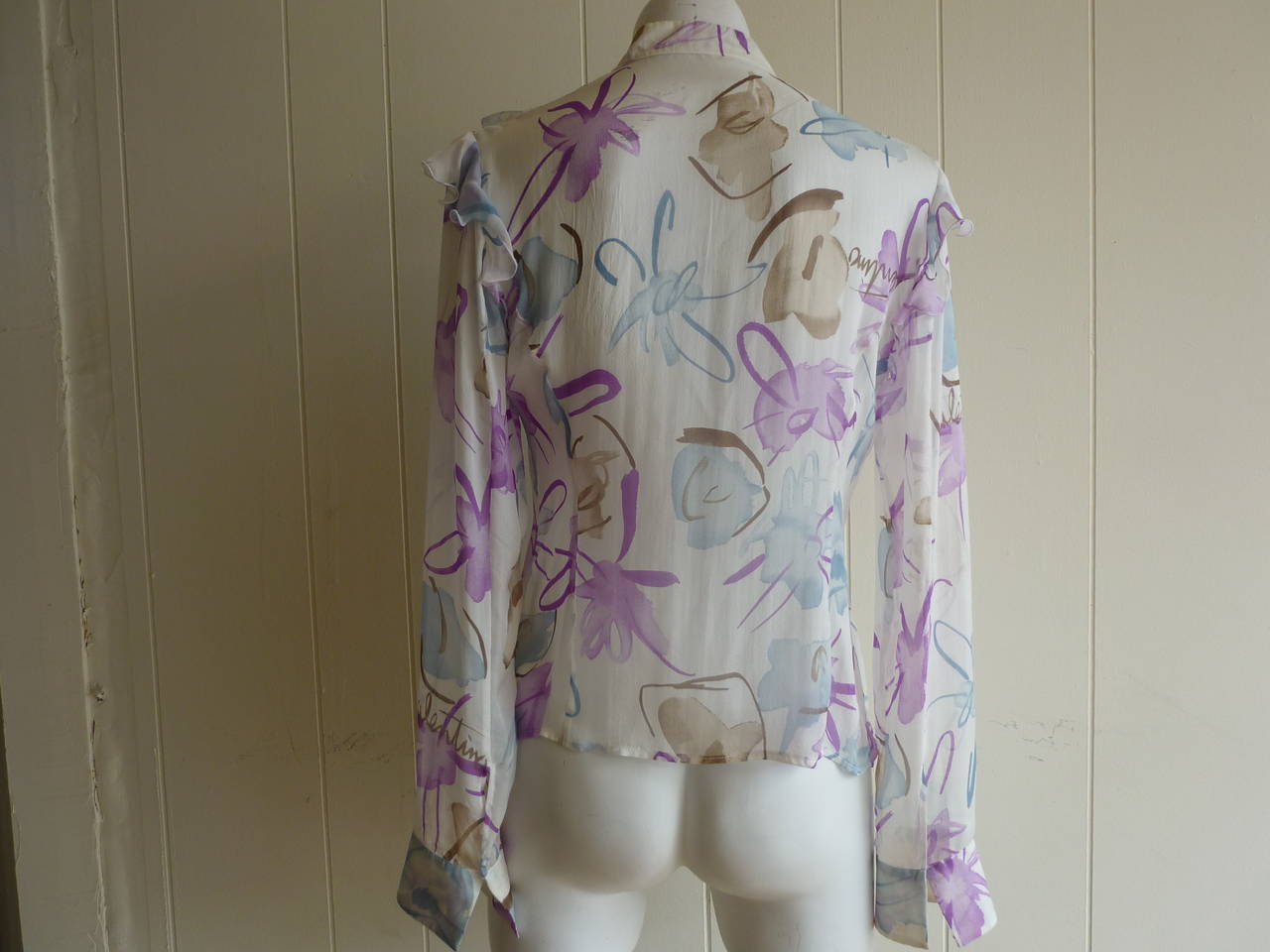 The graphics on this delicate silk blouse are like paint strokes. The background is white with splatters in lilac, teal and browns from bronze to light beige.  There are ruffle on the shoulders and the front of the blouse, and it is signed