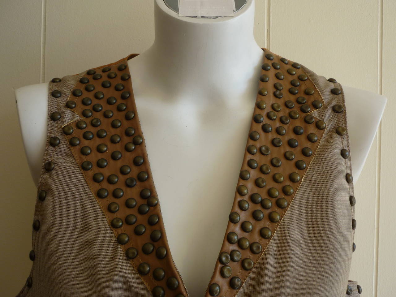 Stunning waistcoat in a fine worsted wool with leather trim at the front v-neckline and pockets. This vest is heavily studded with two slit pockets. The back in a bronze/burnt gold viscose with a half belt and buckle.

Made in Italy
