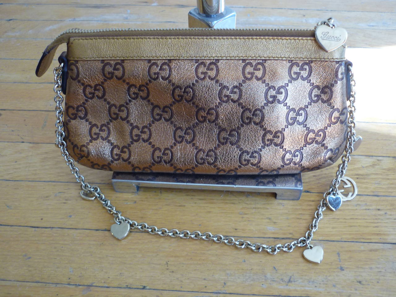 Metallic gold with brown Gucci logo leather and chain link shoulder strap featuring heart charms, this is a lovely evening bag, or go ahead and wear it with jeans!

The interior has a single slit pocket and zip closure at top. It is in excellent