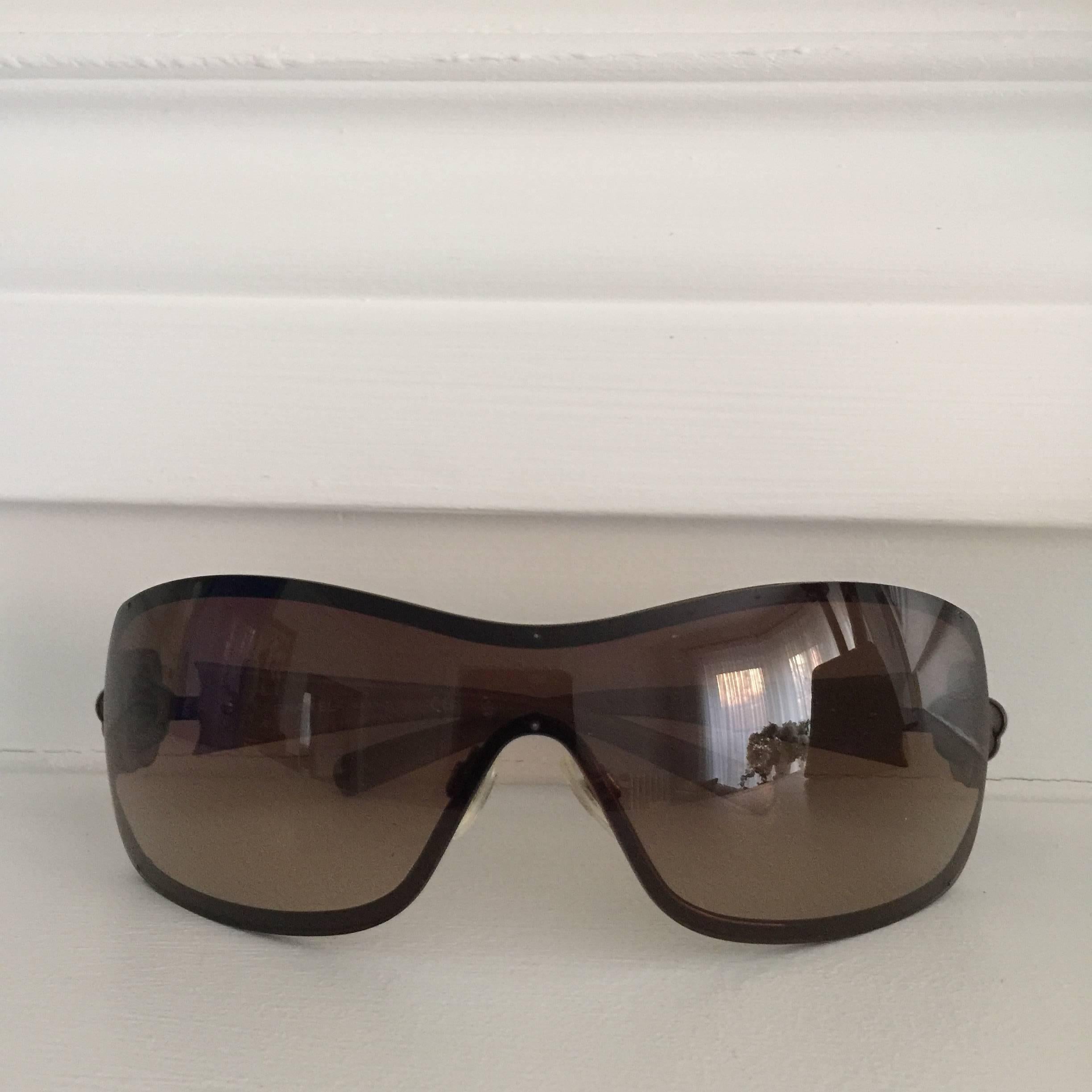 These Ultra-Chic Chanel sunglasses are in one piece 
large lens... in dark amber/mauve.... set amidst a silver frame 
. The arms are wide and feature a Camelia stylized
flower set with Swarovki Crystals and a silver Chanel
logo in the