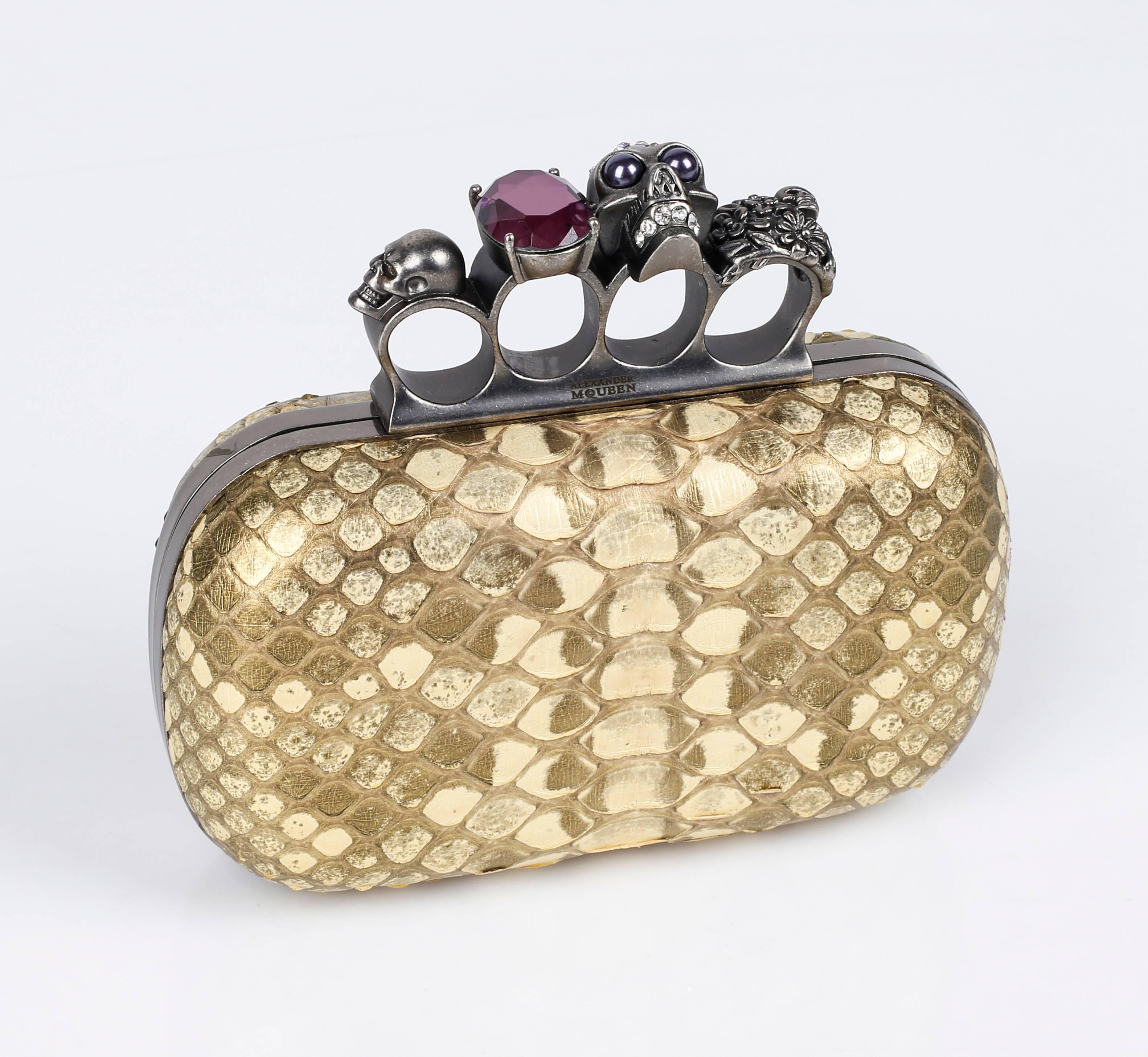 Alexander McQueen Spring/Summer 2010 metallic gold genuine python knuckle duster box clutch. Signature silver-toned hinged clasp with four knuckle rings; embellished in Swarovski crystals, purple gem, and various skulls. Framed hard shell with
