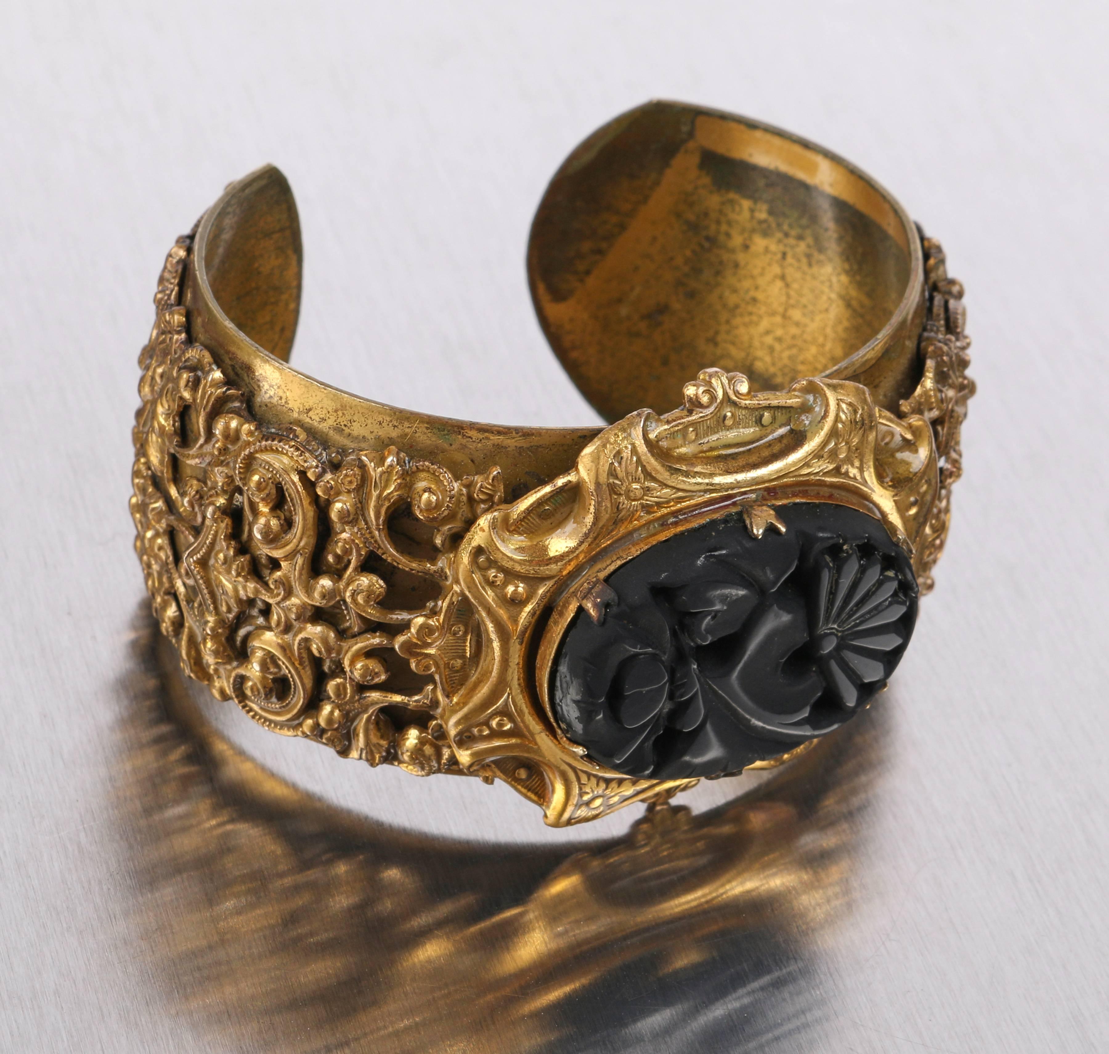 Vintage Victorian Revival c.1930's brass tone metal wide cuff bracelet. Elaborate floral and scroll filigree carved overlay. Large black oval glass stone with floral carved motif set in six prongs. Horizontally oriented stone is set in raised brass
