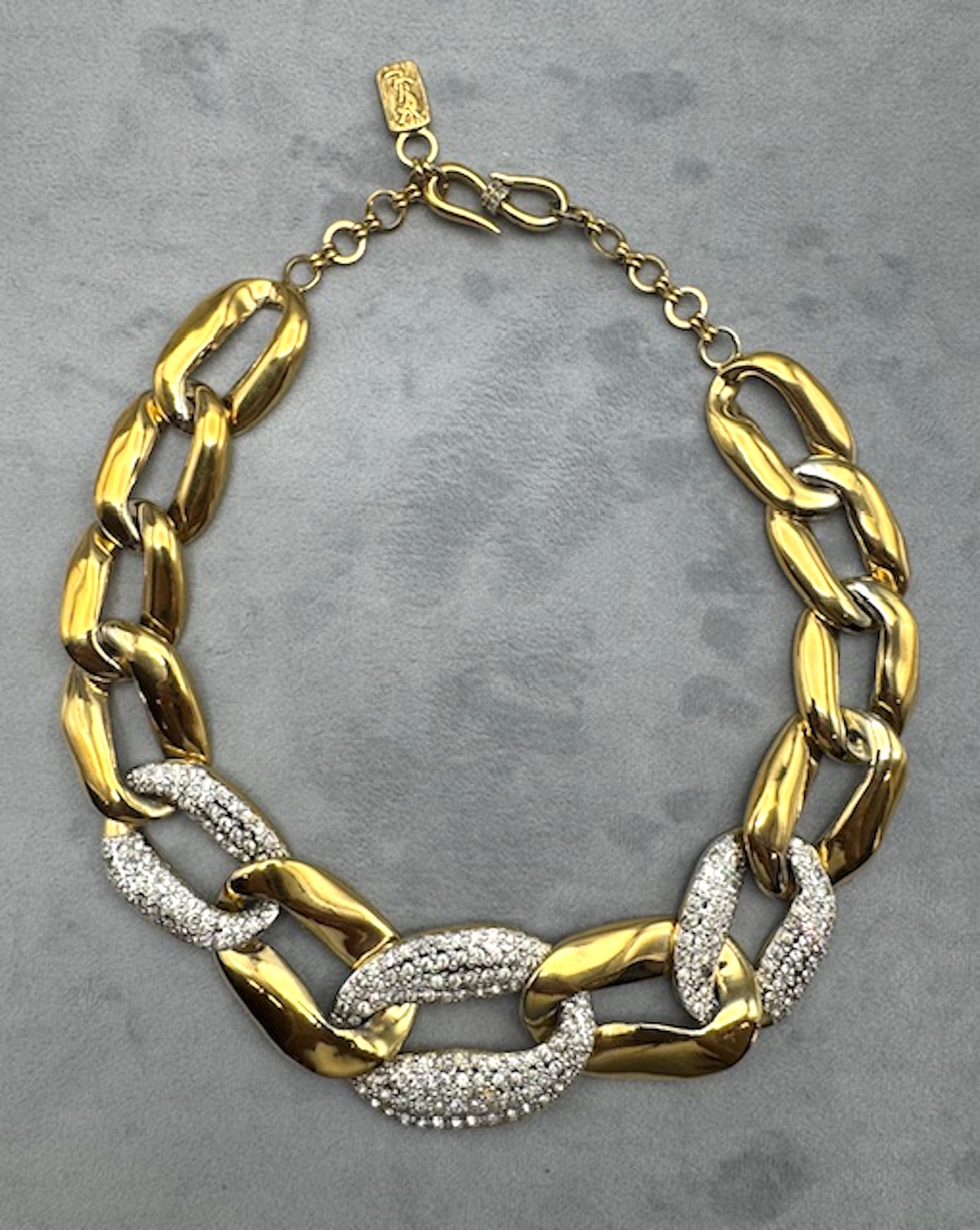 El diseño clásico de los 80 es esta placa dorada  Collar Yves Saint Laurent collar de eslabones grandes.
El eslabón central y más grande mide 5 cm de ancho y está engastado con brillantes. Desde el eslabón central de brillantes, cada cadena se va