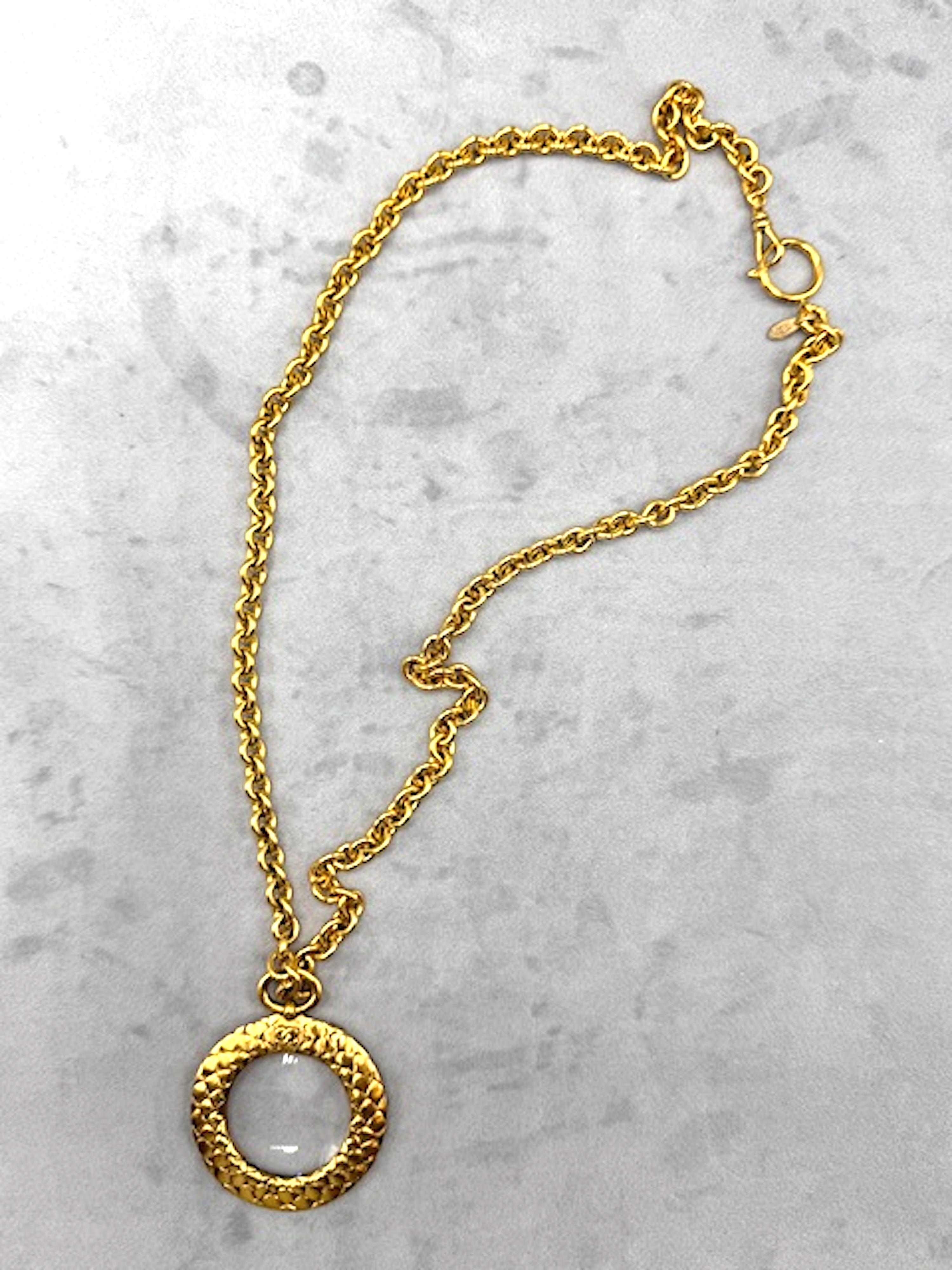 Un icónico collar colgante de lupa con marco acolchado de Chanel de la primavera de 2002. El colgante tiene una lente de aumento de cristal engastada en el marco redondo acolchado y mide 5 cm de diámetro. La altura del colgante con el anillo es de 2