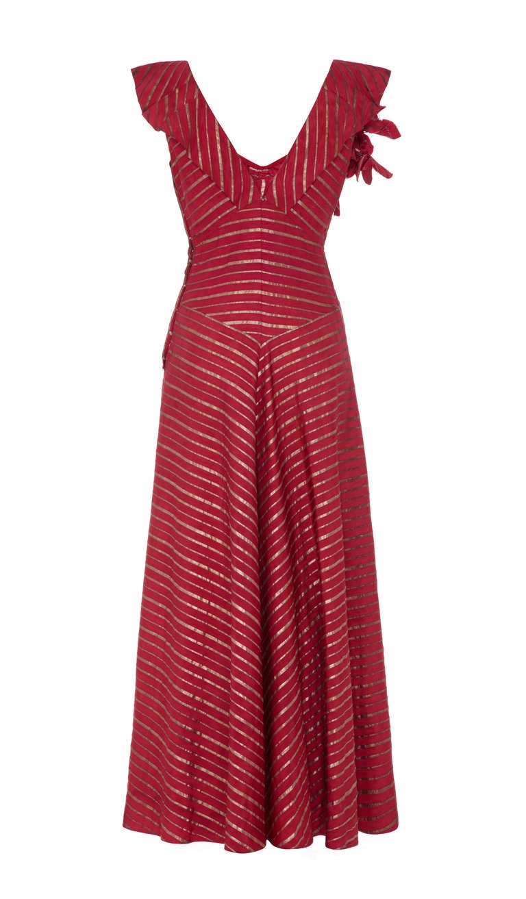 Gorgeous maroon silk gown with stripes of metallic gold lame. This dress is full length and features ruffle shoulders, a deep V neckline and front gathering with a huge floral corsage in the same fabric. It fastens with poppers to the side and is in