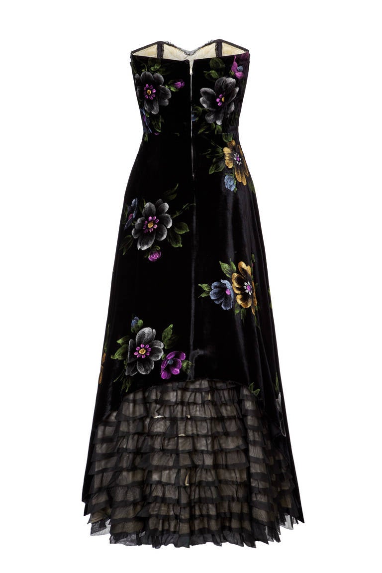 Sensational black velvet gown with beautiful hand painted floral motifs in subtle hews of purple and green.  This gorgeous full length, strapless evening dress features pretty black net ruffles across the bust and to the back of the skirt. It