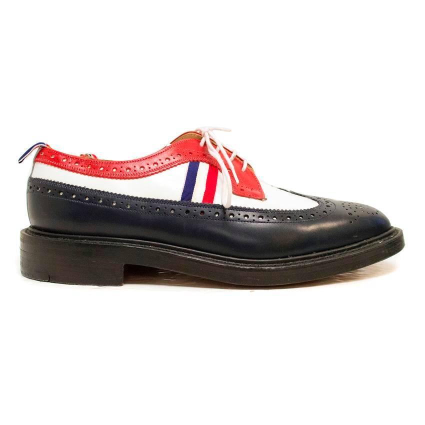 Thom Browne navy blue, white and red leather panelled brogues with a blue, white and red stripe down the middle of the shoe. There is a blue, white and red pull at the back of the shoe, and the shoes have white laces with black midsoles.

Slight
