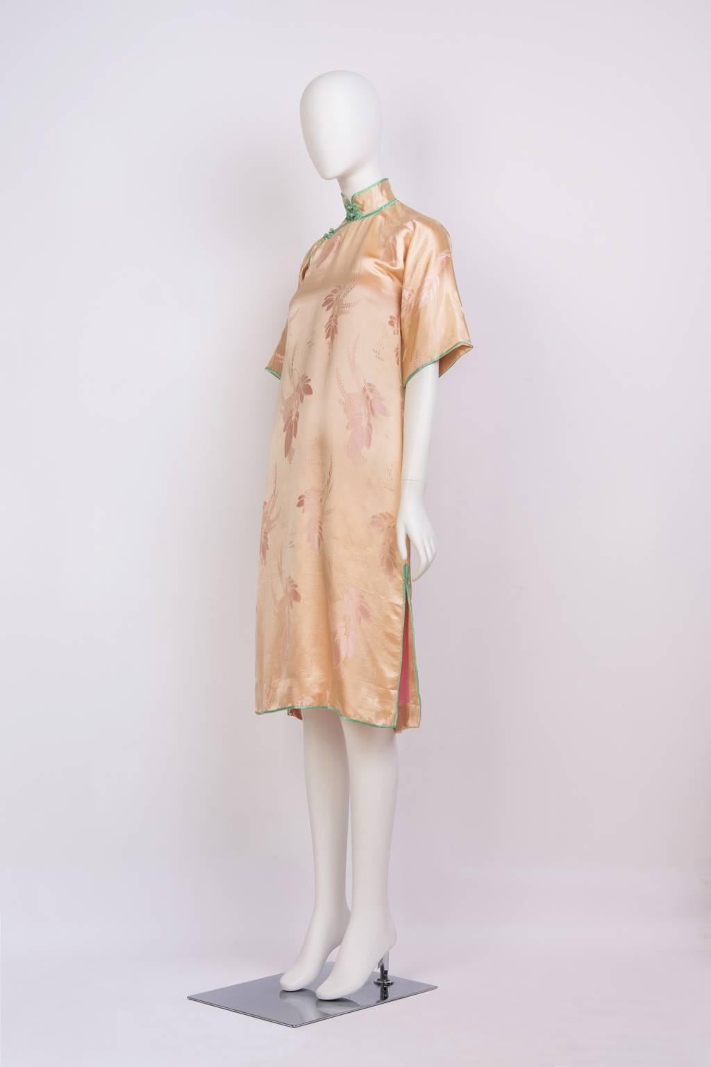 Short sleeve, knee-length cheongsam with side slits and frog closures in peach silk floral jacquard with green trim.