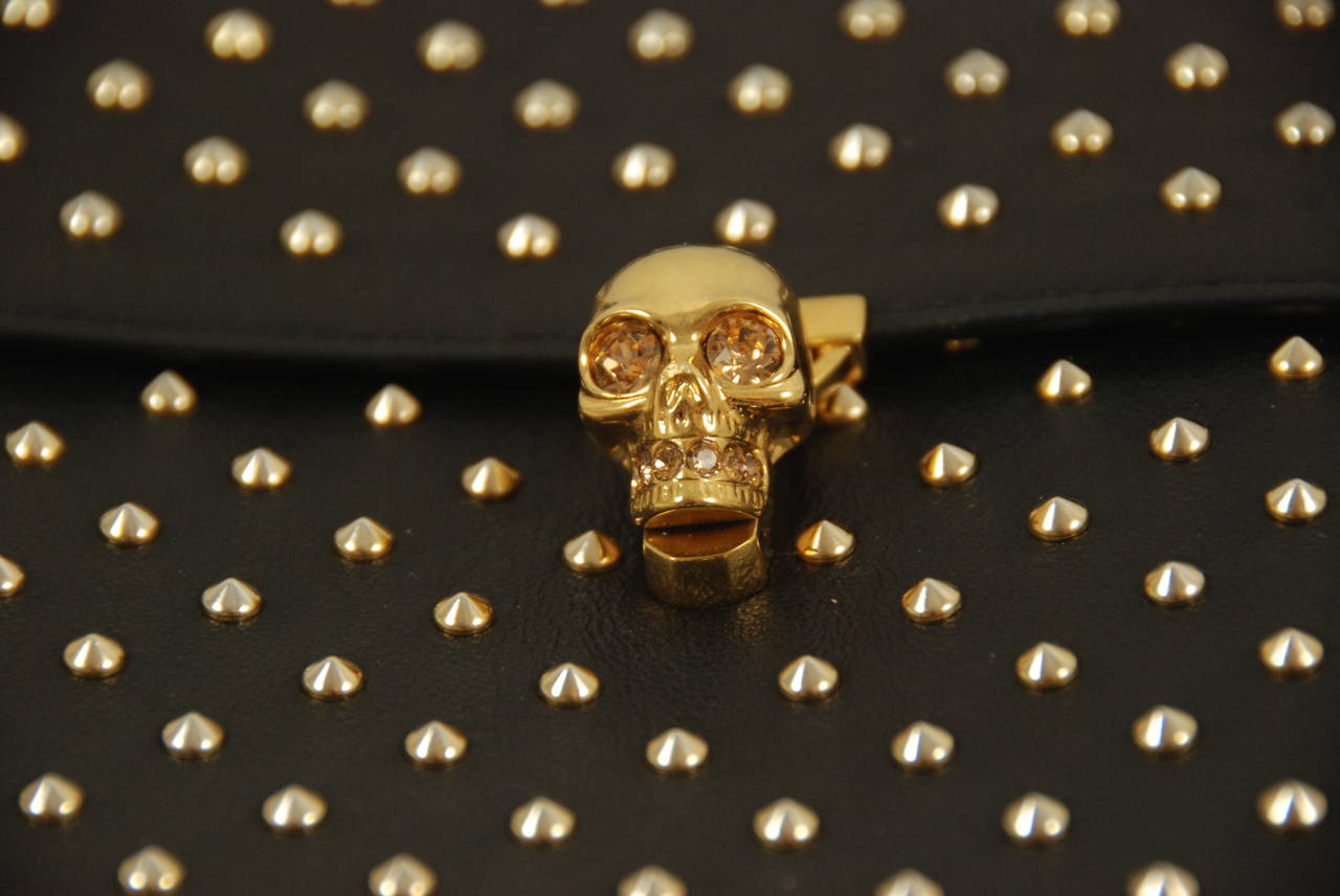 Alexander McQueen black leather envelope style clutch with brass studs and skull clasp. The skull has rhinestone eyes and embellishments. The rhinestones are yellow. Clasp opens by twisting either right or left.  Clasp works well and bag closes