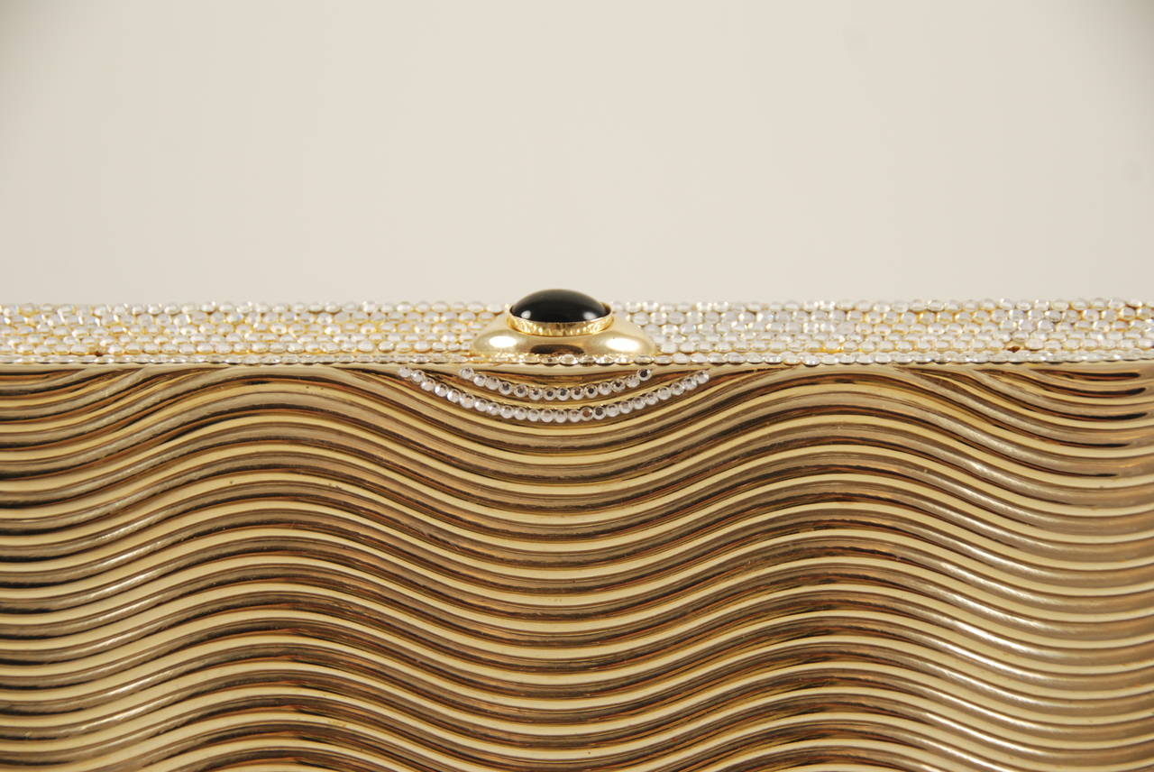 Judith Leiber minaudiere circa 1990s. This one is both elegant and sophisticated. The top, sides and bottom are encrusted with clear rhinestones while the front and back are gold metal with a wave pattern  to the metal. The clasp is a cabochon black
