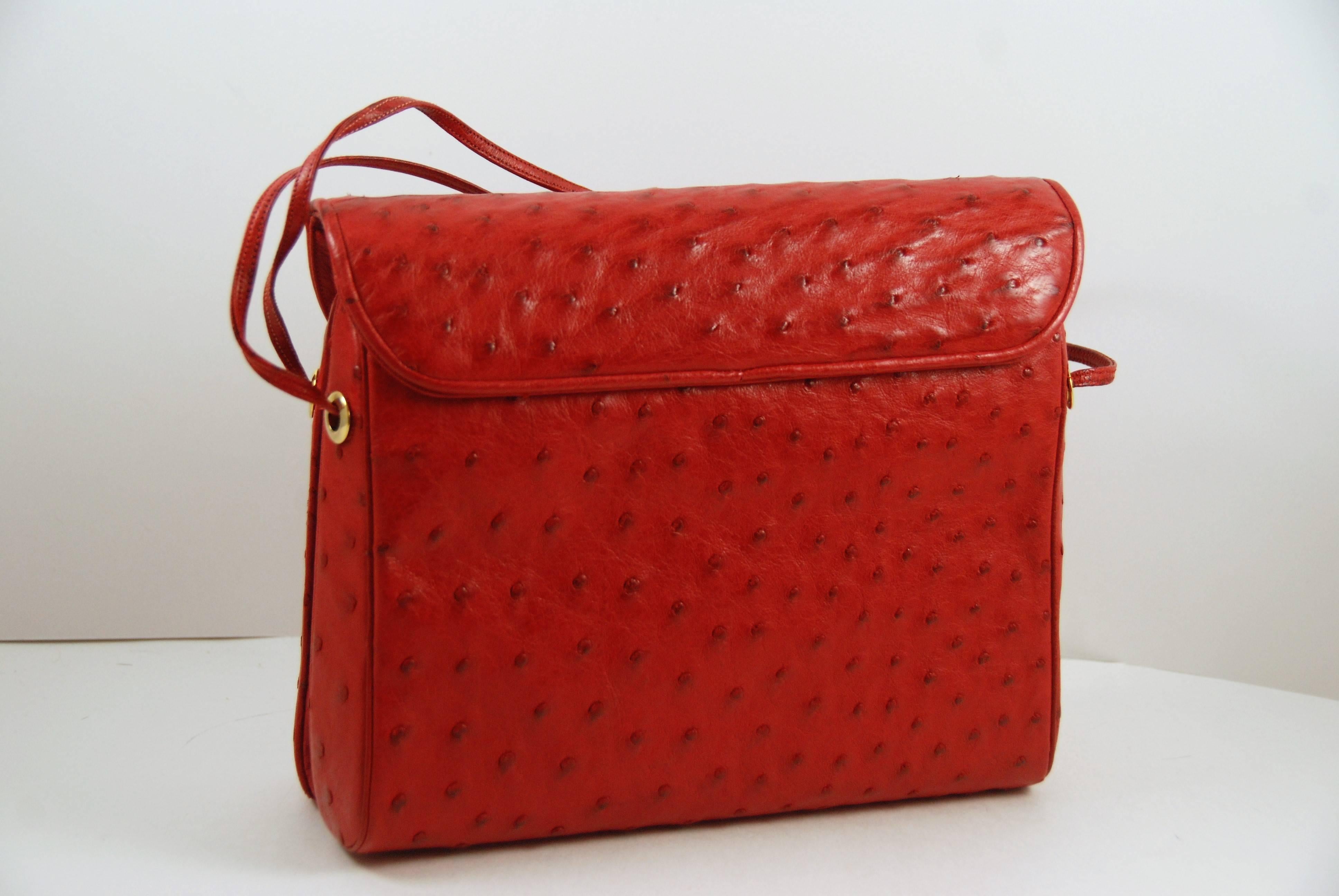 Judith Leiber shoulder bag is done in vibrant geranium red ostrich and features a flap top with magnetic snap closure. It has a double red ostrich shoulder strap and gold tone hardware. The interior is done in red canvas and has one large main