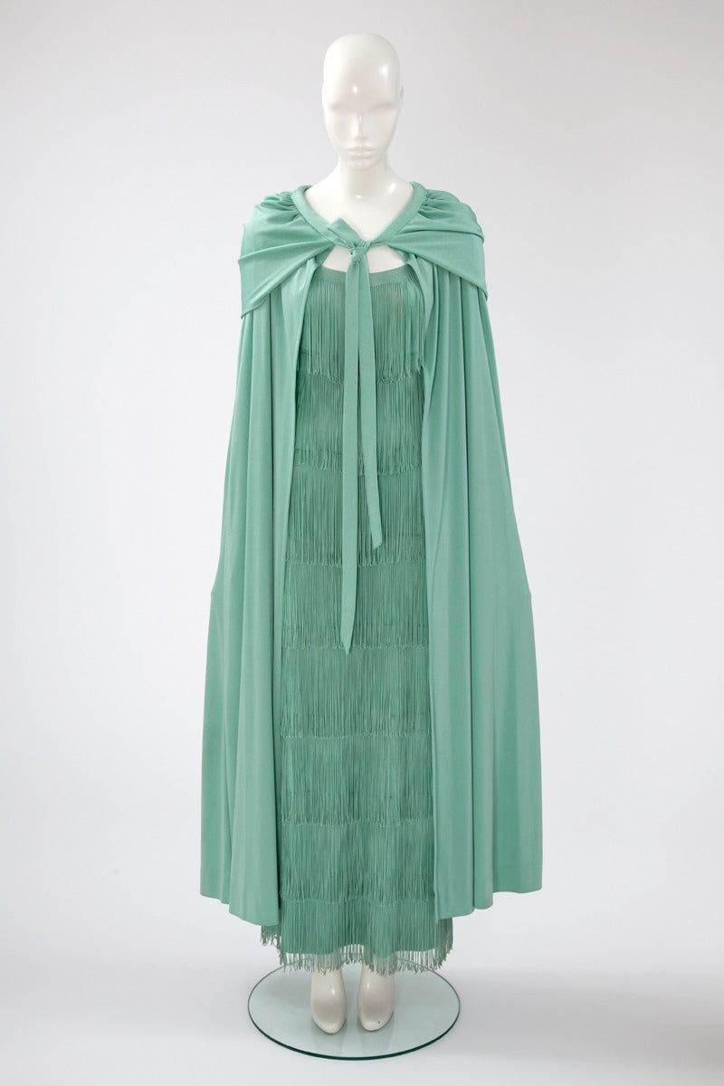 Quintessential example of the legendary 70's decade, this Loris Azzaro fringed evening gown and matching cape will create sensation ! Constructed in jade green jersey, this columnar gown is adorned with multiple fringed tiers, while the full length