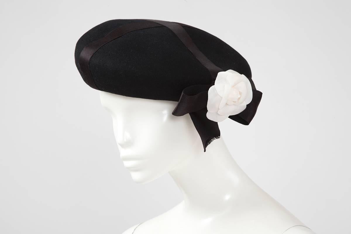 This vintage Chanel beret will provide any outfit an effortlessly chic spin. Cut from soft wool-felt, this warm and cozy hat features black silk satin ribbons & the recognizable iconic Chanel camellia on top of a bow. It is a size 56 and is signed