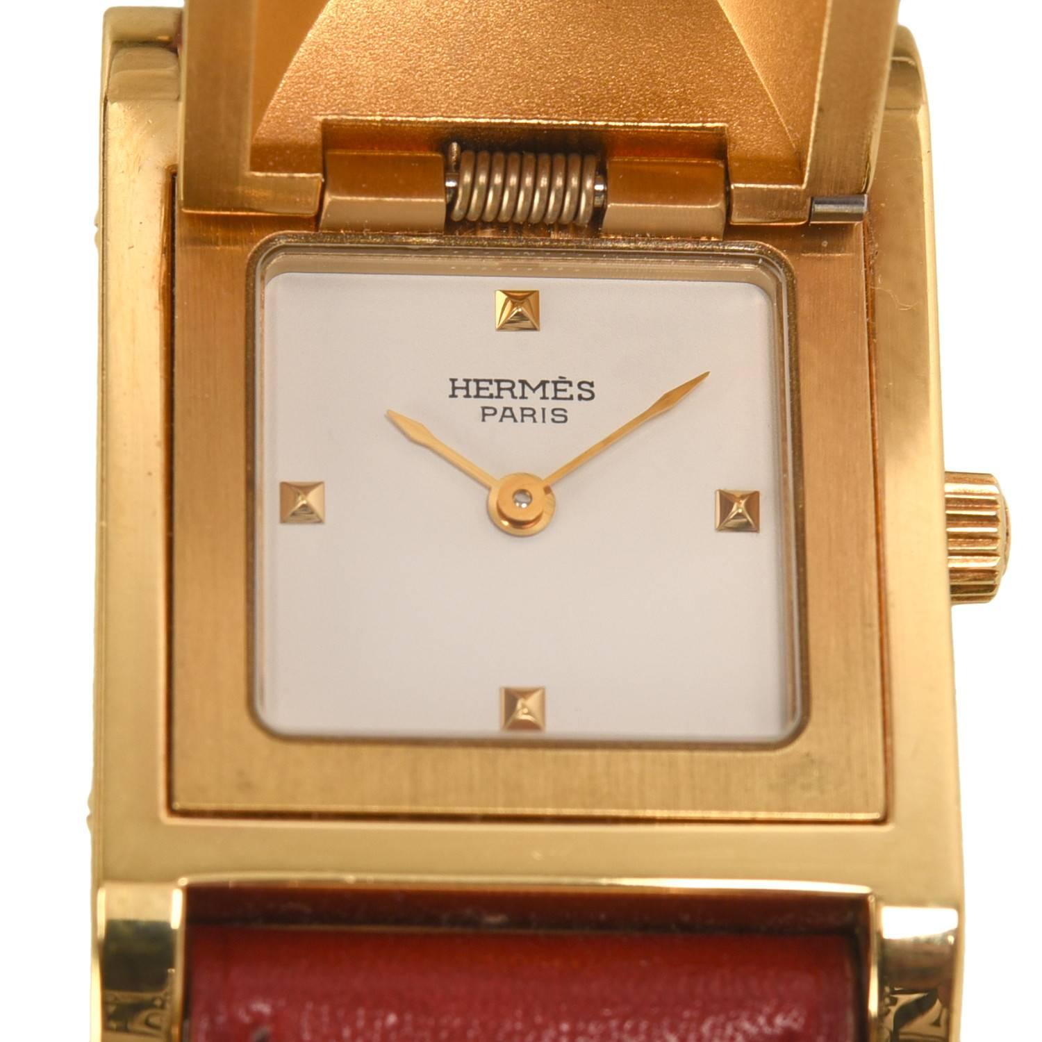Hermes Medor watch size PM with rouge calfskin leather band.

This watch is made of gold plated stainless steel accented with pyramid studs, the face being hidden under the center stud. The calfskin leather band is adjustable and has a Hermes