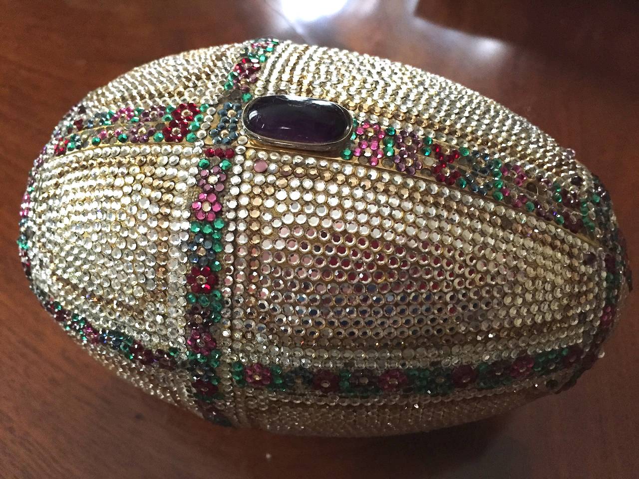 Delightful Judith Leiber Faberge Style Egg Minaudiere

Designed as an egg with a trellis of tiny flowers in amethyst , emerald and ruby.
Between the trellis is clear crystal with a topaz cartouche inside.
The missing crystals are mostly the
