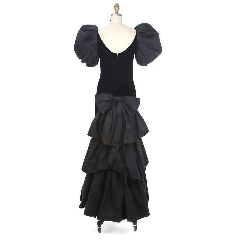 This is a haute couture gown in black velvet by Yves Saint Laurent, circa 1980s. It features a puffed short sleeve and a volumunus tiered train.  Other details include a detachable bow at the back waist, high neckline with a scooped back, and a