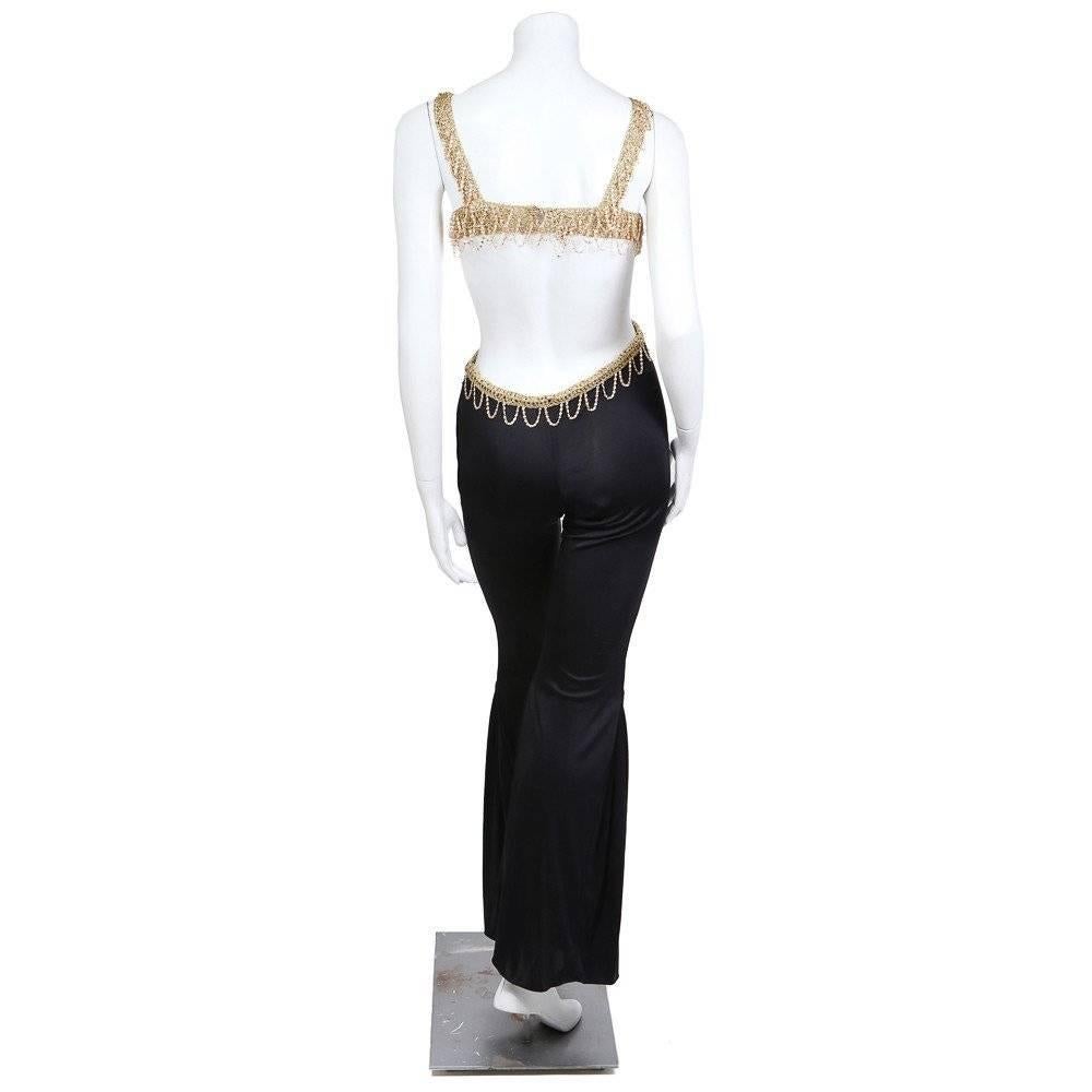 This is a jumpsuit circa the 1970s.  The top is knit with gold thread and features hanging looped gold chains to create a fringe.  These same looped fringe chains also go around the waist, eluding to a belt detail. The leg is a wide flare starting