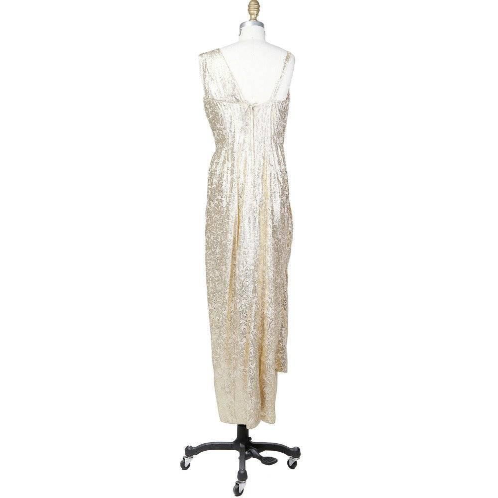 This is a custom gown by Blackwell, c. 1960s.  It is a made from a shimmering gold lightweight brocade.  It features a long front drape that starts able the waist just under the bust.  The shoulder straps are also different sizes, one spaghetti and