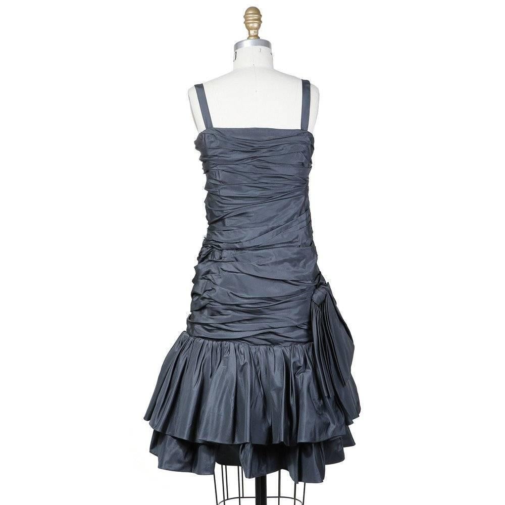 This is an haute couture dress by Yves Saint Laurent c. 1980s.  It is made from charcoal colored satin. It features gathered and folded fabric all around with a two tiered ruffled skirt. Other details include a bow at the side and half inch wide