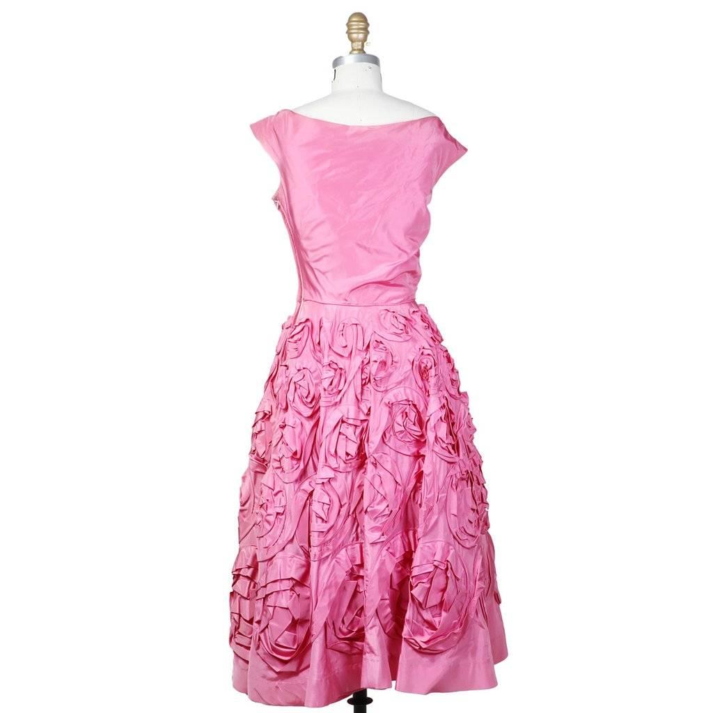 This is a pink satin dress by Ceil Chapman c. 1950s.  It features a hanging drape over the neckline and 3D textured rose details around the skirt.  Some more rose detail can be seen at the neckline just behind the drape.  The dress includes a tulle