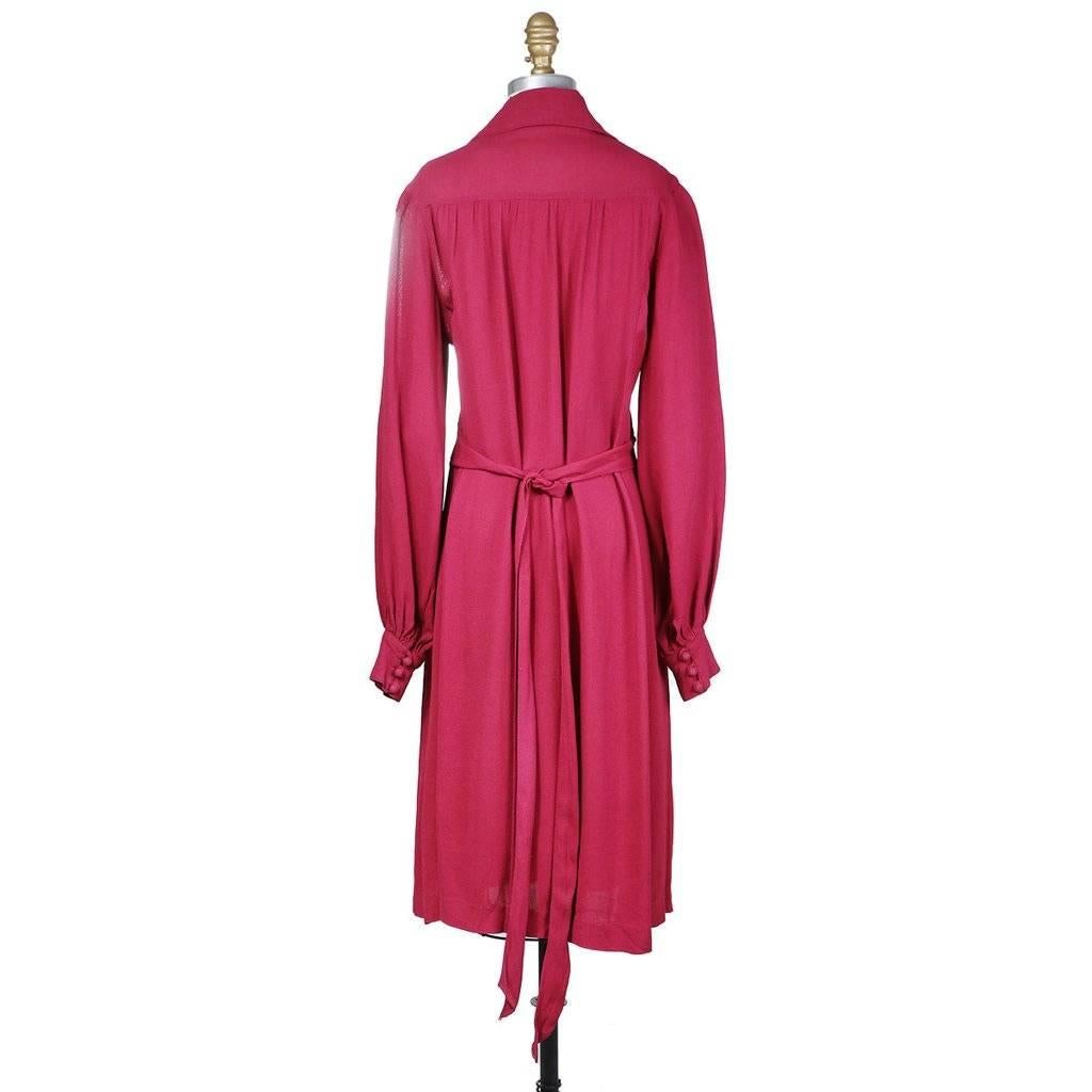 This is a magenta crepe dress from Ossie Clark c. 1970s.  It features long bishop style sleeves with large cuffs as well as a large collar. The front center button closures begin at the waist and end at the neckline.  There is also an attached waist