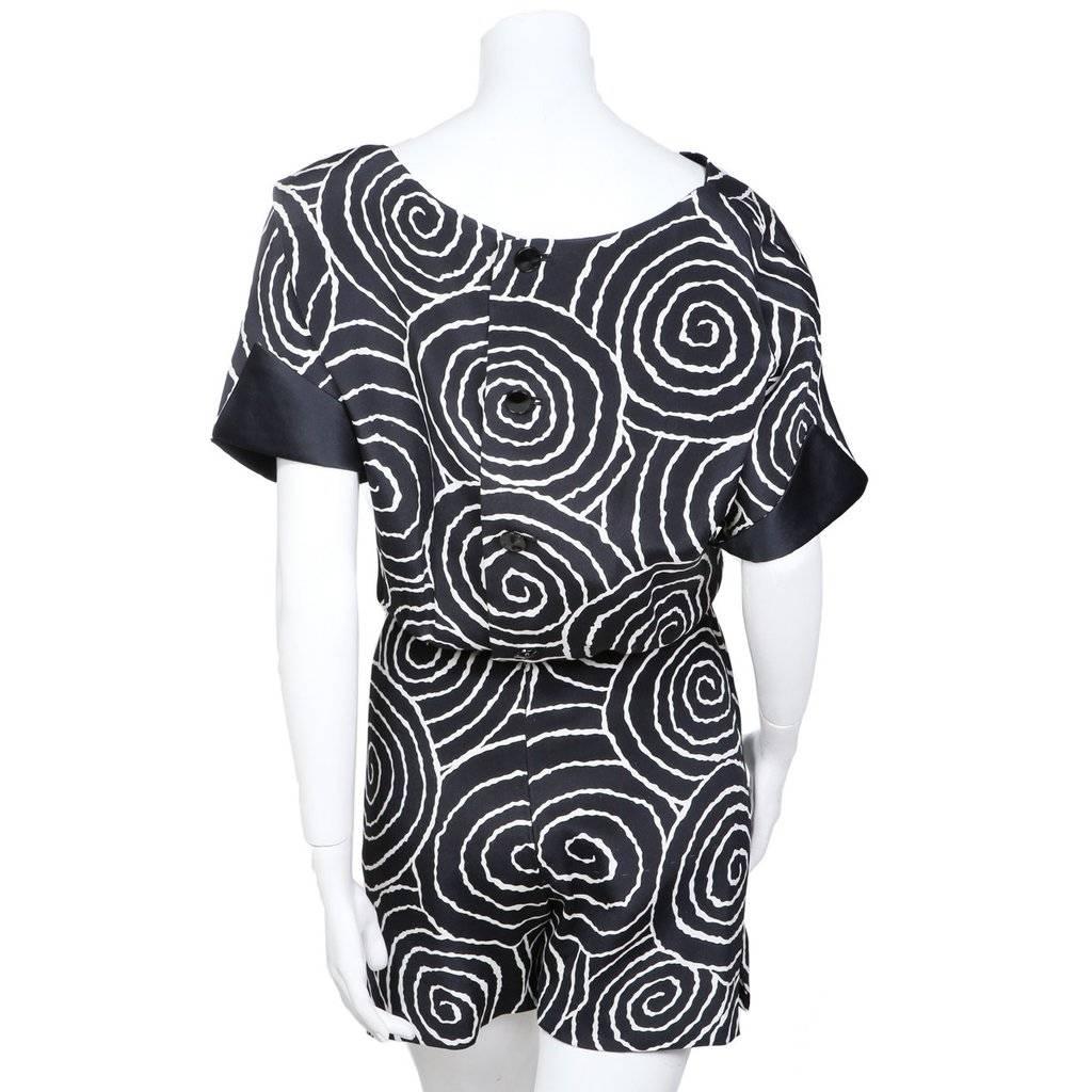 This is a black and white romper by James Galanos c. 1980s.  It features an optical spiral print, fold over short sleeves that reveal black silk satin lining, and slits on sides of pant legs.  The romper includes an elastic waist and buttons down