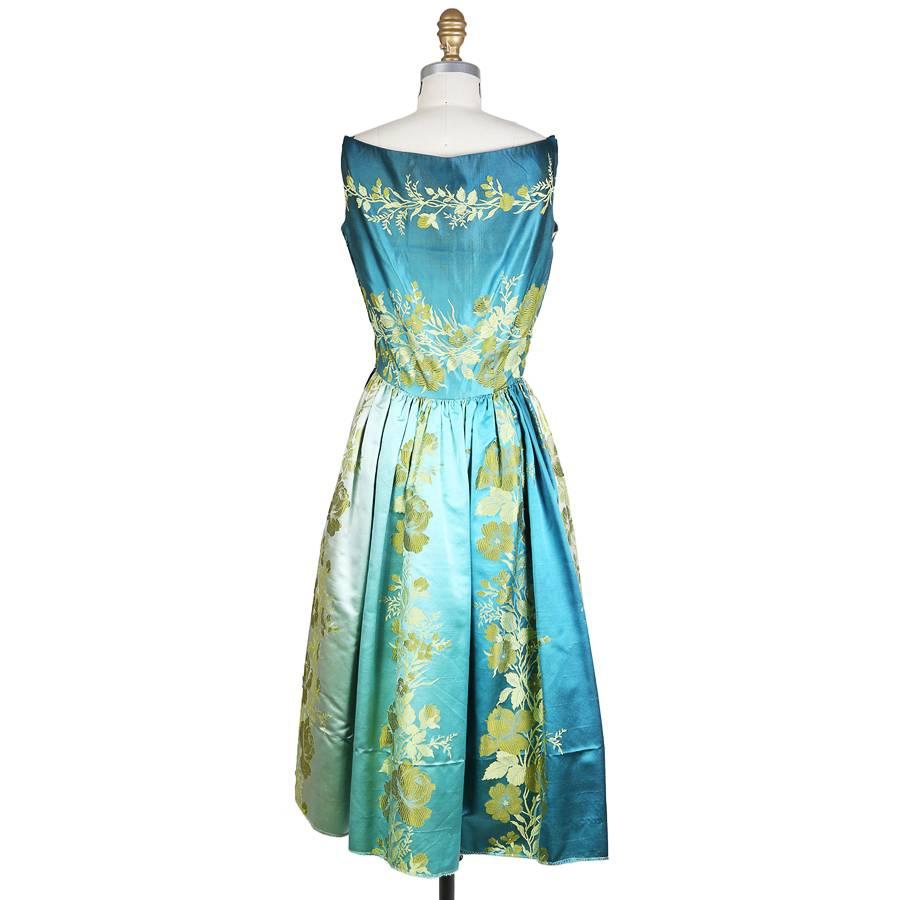 This is a Burke Amey dress c. 1960s.  It is made from a satin brocade that features a subtle ombre and floral motif.  The dress has a boat neckline with short shoulder straps that attach by hook and eye.  The closure is a hidden side zipper with
