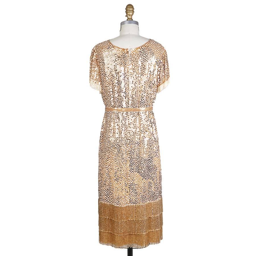 This is a shift dress by Norman Norell c. 1960s.  It is made with a cream colored satin base which is covered in gold paillettes.  The arms and bottom of the dress feature a beaded fringe.  It includes a matching sequin belt.  The closure is a