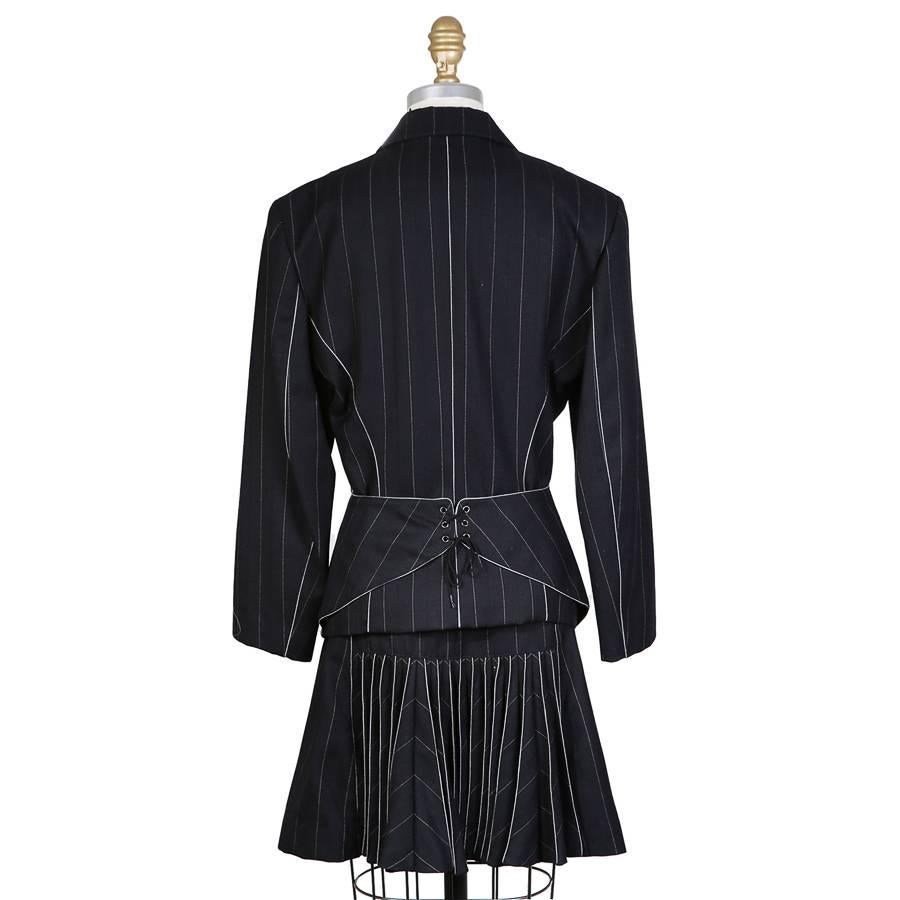 This is a navy blue pin stripe skirt suit by Azzedine Alaia c. 1990s.  It features white piping details to create style lines and has a single column of buttons on the front jacket closure. The back features a corset detail on the waist of the