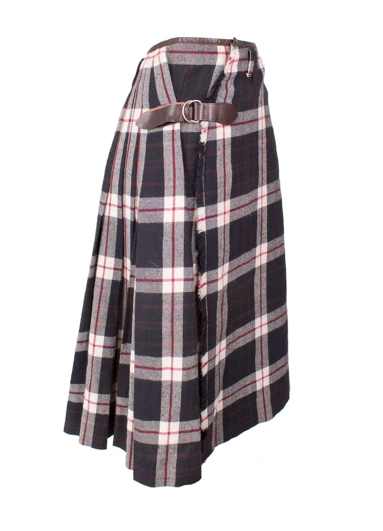 This is a wool kilt by Jean Paul Gaultier c. 1990s.  It features a red white and navy plaid with brown leather trimming.  The back is pleated and there is frayed edges in front.  The skirt wraps around and closes with buttons, hook and latch, and a