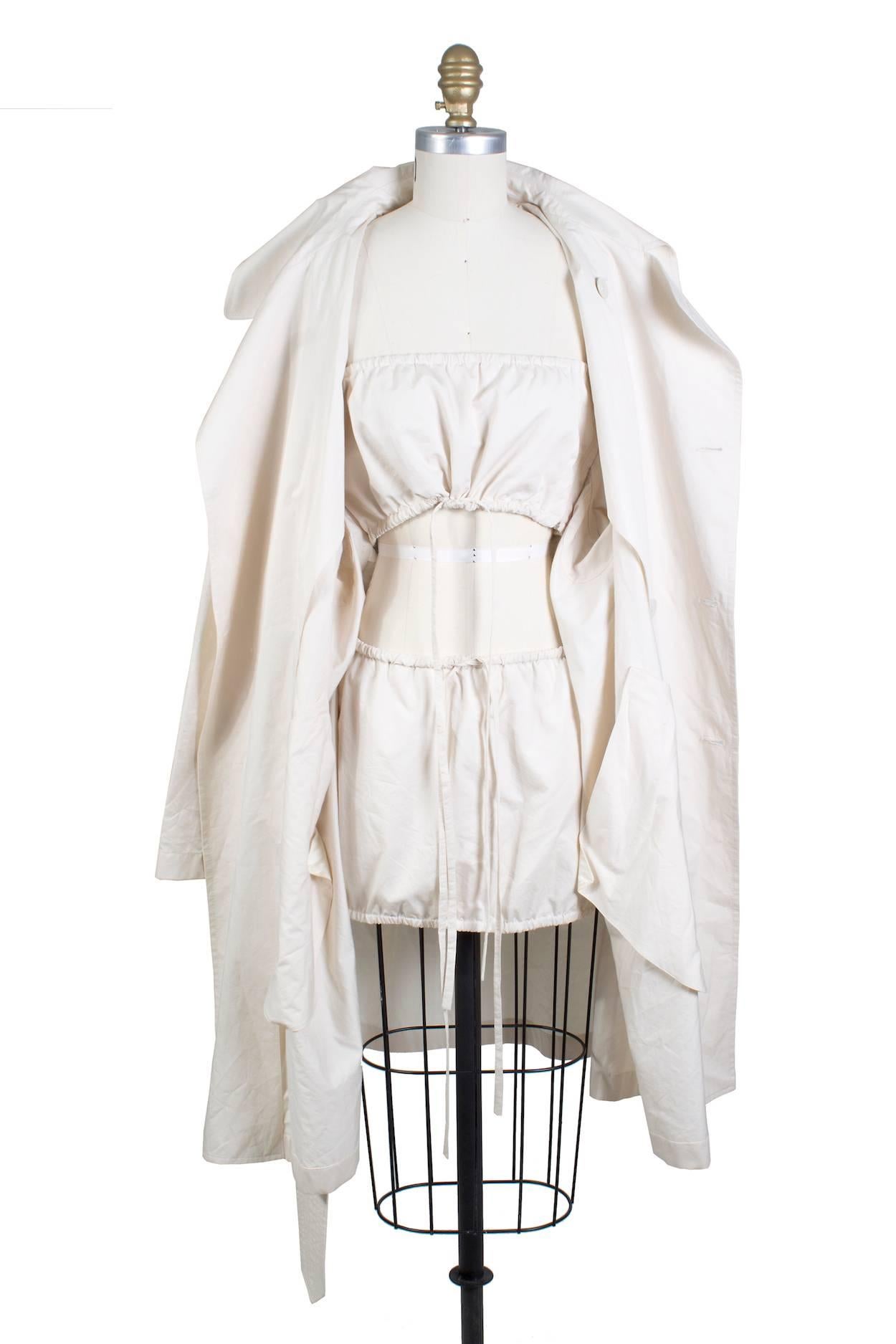 This is a beige trench coat by Jean Paul Gaultier c. 1990s.  It has an oversized fit and features an attached inner bando top and mini skirt (which both have elastic drawstring and zippers). The jacket is double breasted with irridescent buttons.