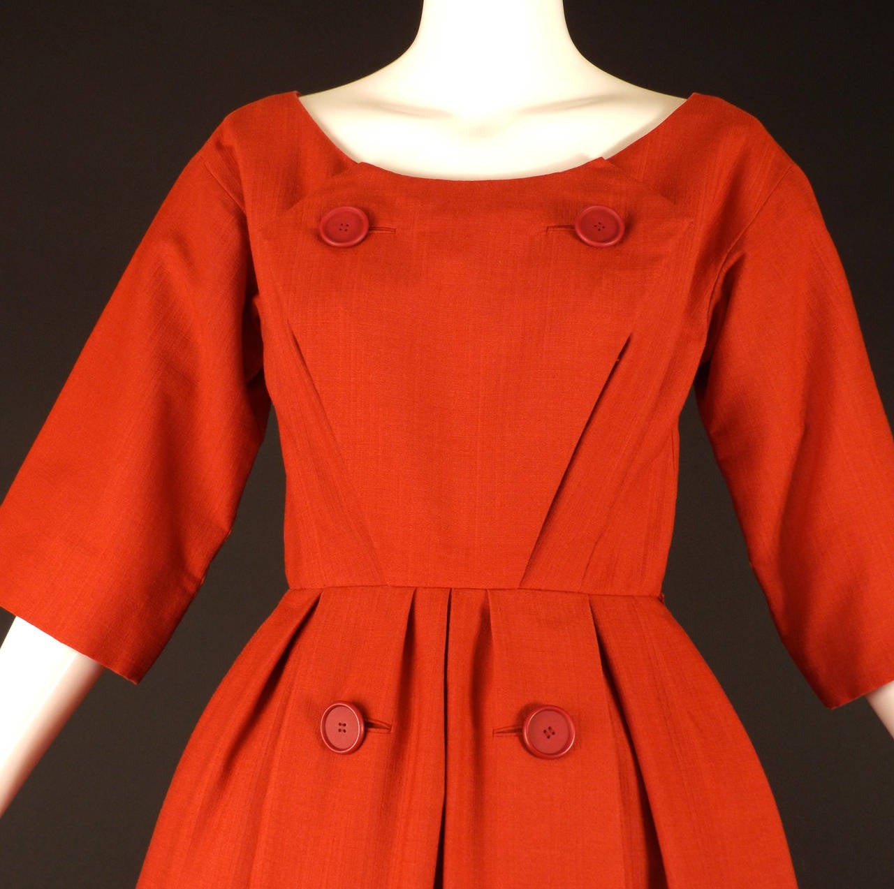 Adorable c.1958 silk and wool blend dress in a brick red. The dress has a scoop neckline with a bib front panel down the front with two large red button decoration on the chest. Single released pleats point upward from the waistline to the bust for