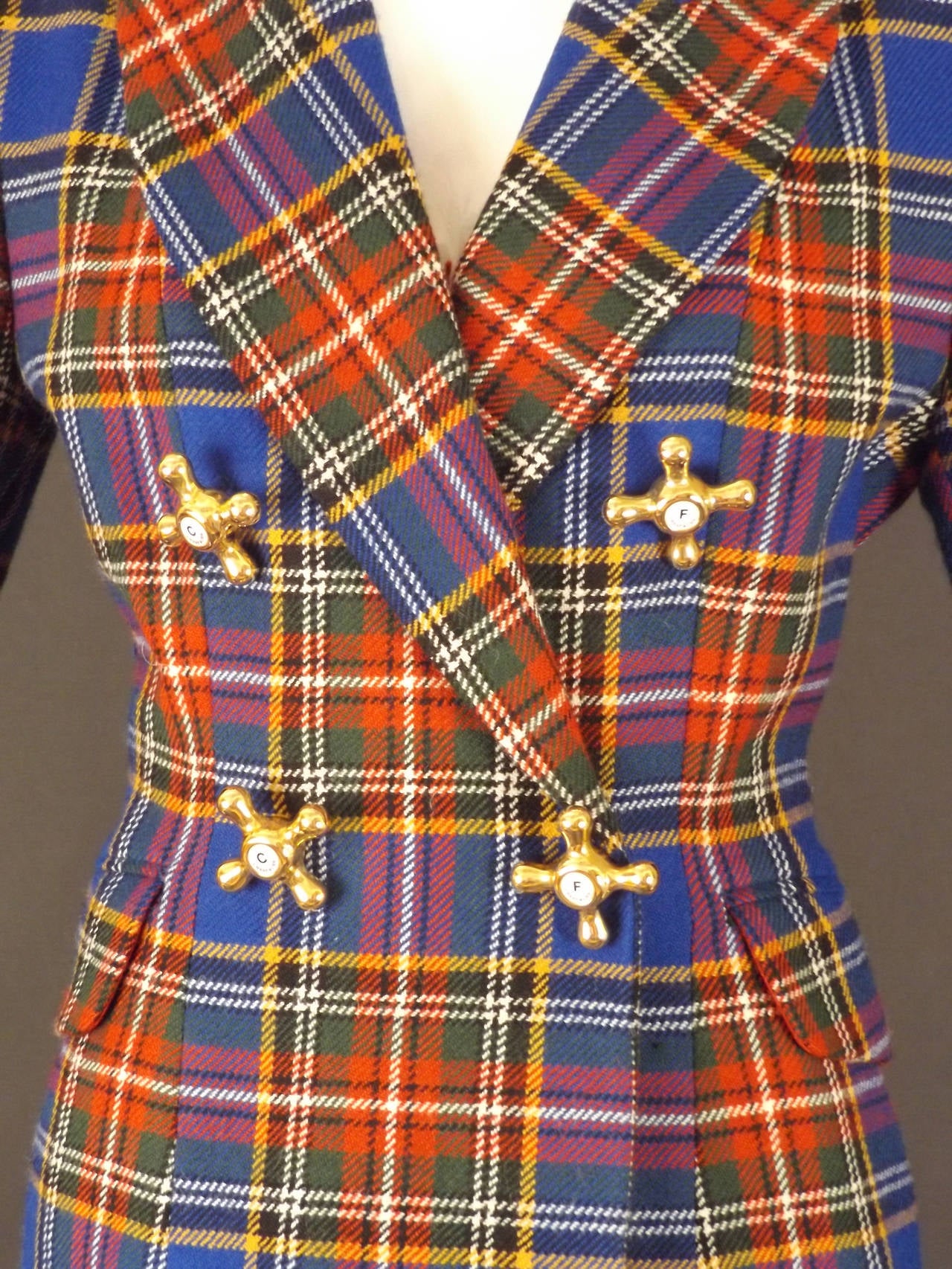 Fall, 1991 Collection. The suit is a blue, red, white, green and yellow wool plaid. Small collar and wide notched lapels.  Double breasted with snap and button closures hidden beneath the gold and white faucet button decorations. The faucets are