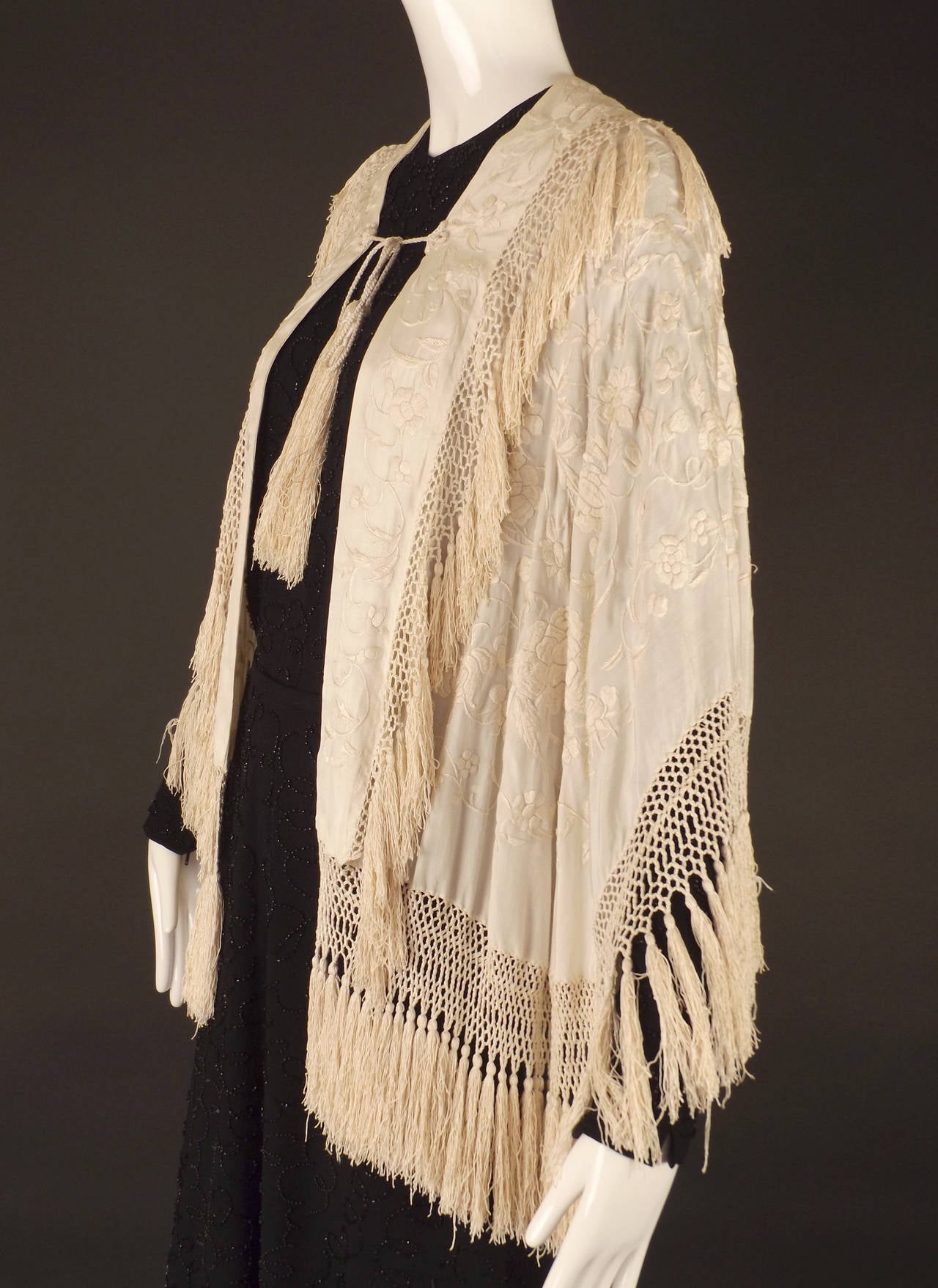 Gorgeous c.1920 cape made from an ivory silk embroidered piano shawl. The piano shawl has a hand embroidered floral pattern through out and trimmed with long, hand tied fringe.  Cord ties at the front chest with a long ivory tassels at the bottom.