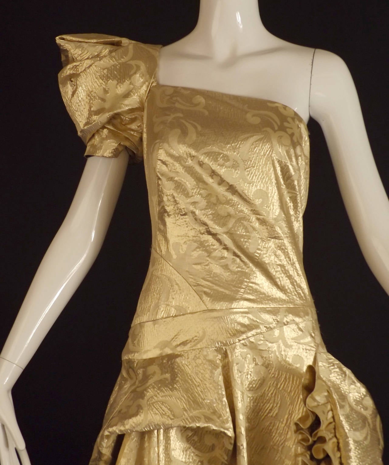 Stunning 1980s ball gown in a gold tissue lamé brocade.  One shoulder bodice with a pleated pouf short sleeve on the right arm.  Interesting seaming through the bodice to the dropped waistline. The skirt is in yards of fabric  that is tucked,