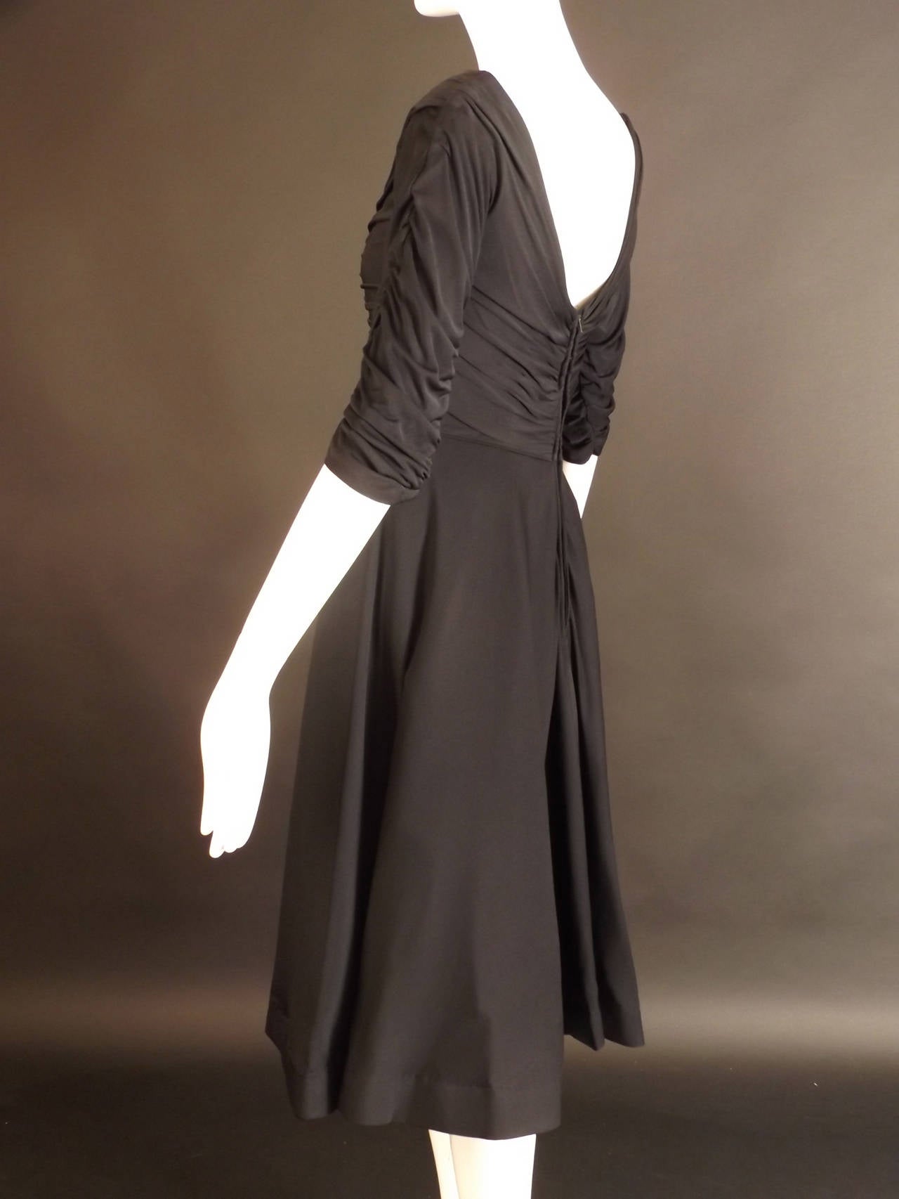 Gorgeous 1950s dinner dress in a black silk jersey knit. The dress has a scoop neckline with shirring on the shoulders and down the side seams, draping the fabric across the body. 3/4 Set in sleeves are also shirred down the seams.  The skirt falls
