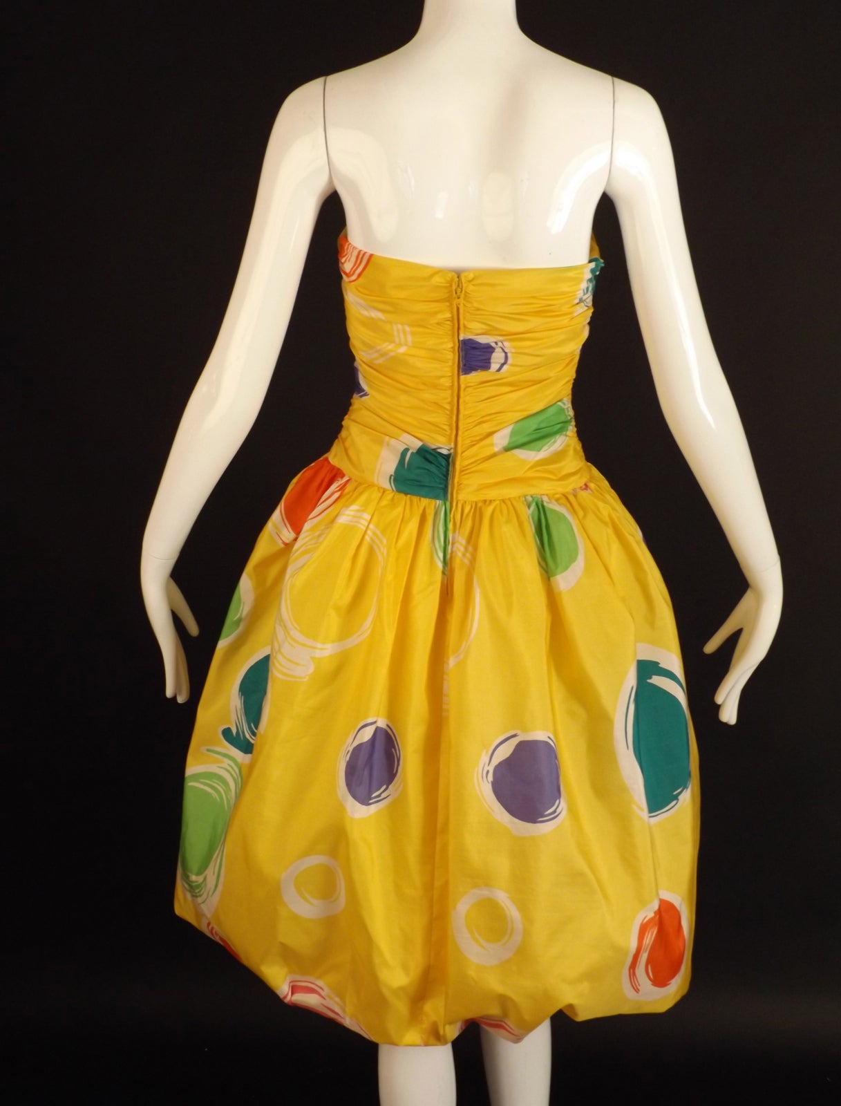 Adorable 1980s cocktail dress in yellow silk with a purple, green, orange and white polka dot pattern. The dress has a bustier liner with the silk shirred down the sides creating draping across the front and back. Dropped waistline with a full